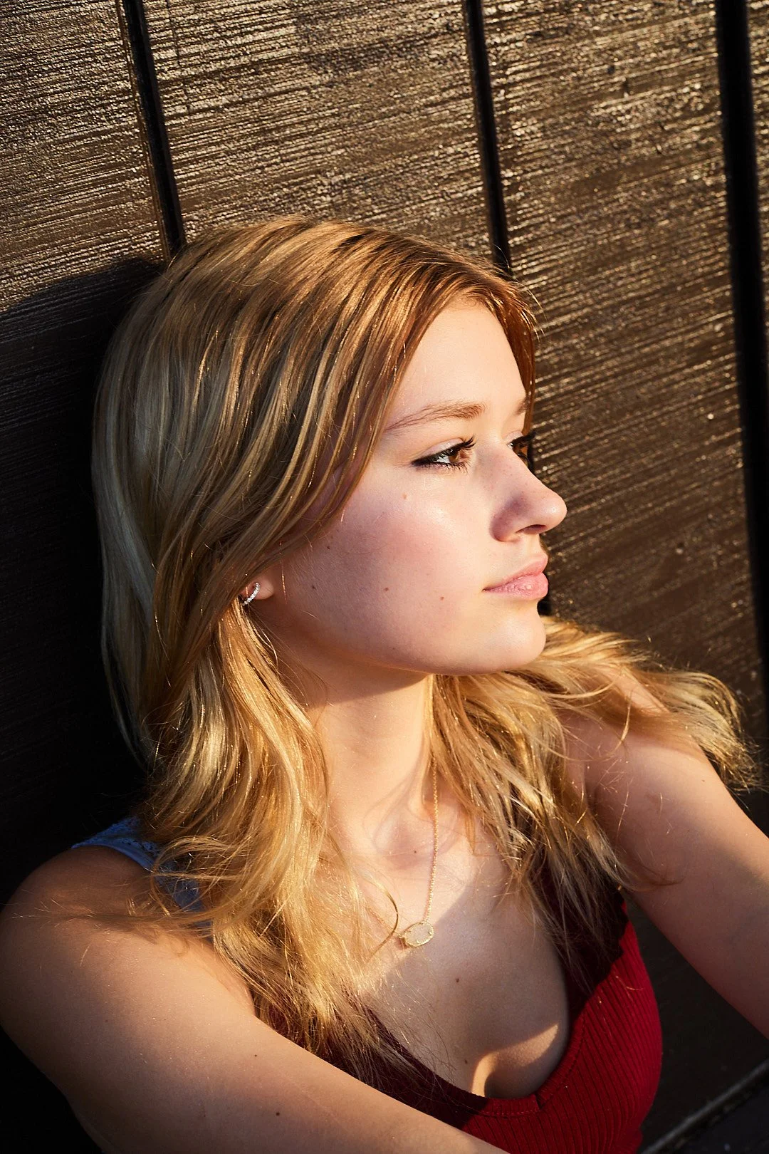 Young woman with blonde hair looking sideways, wearing a red top and necklace, sitting against a wooden wall.