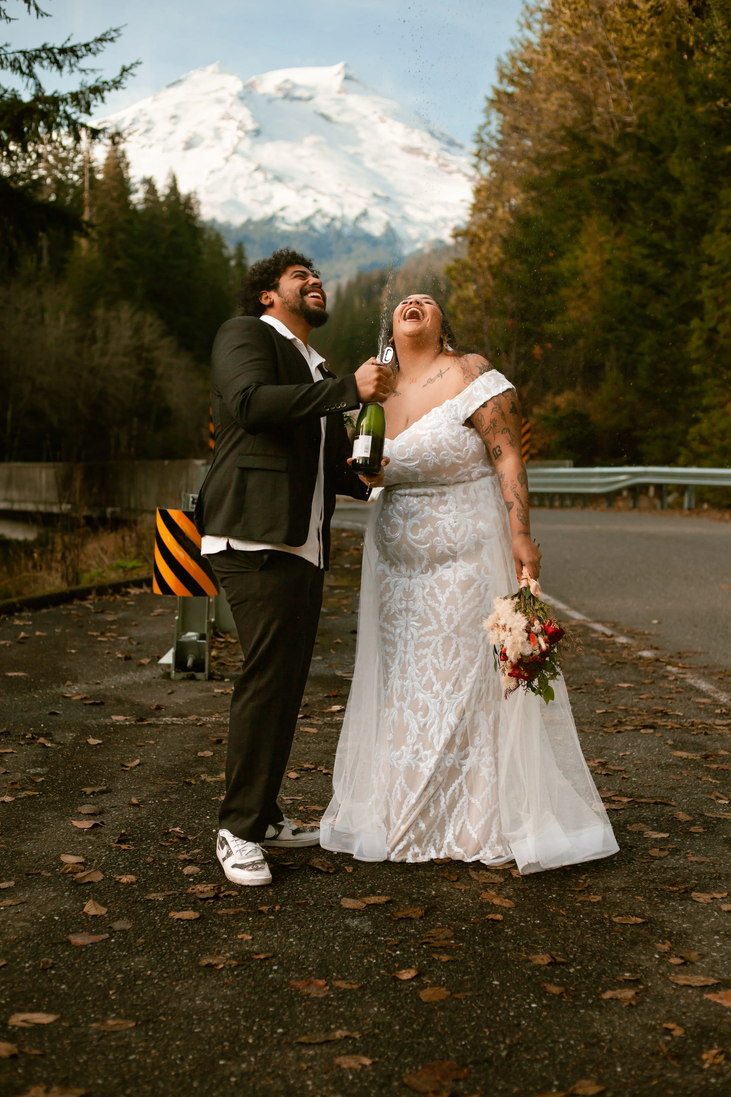A newlywed couple celebrating outdoors on a mountain road with snow-capped peaks in the background. The bride is in a white lace wedding dress, holding a bouquet, and laughing while the groom in a black suit and sneakers is opening a bottle of champa