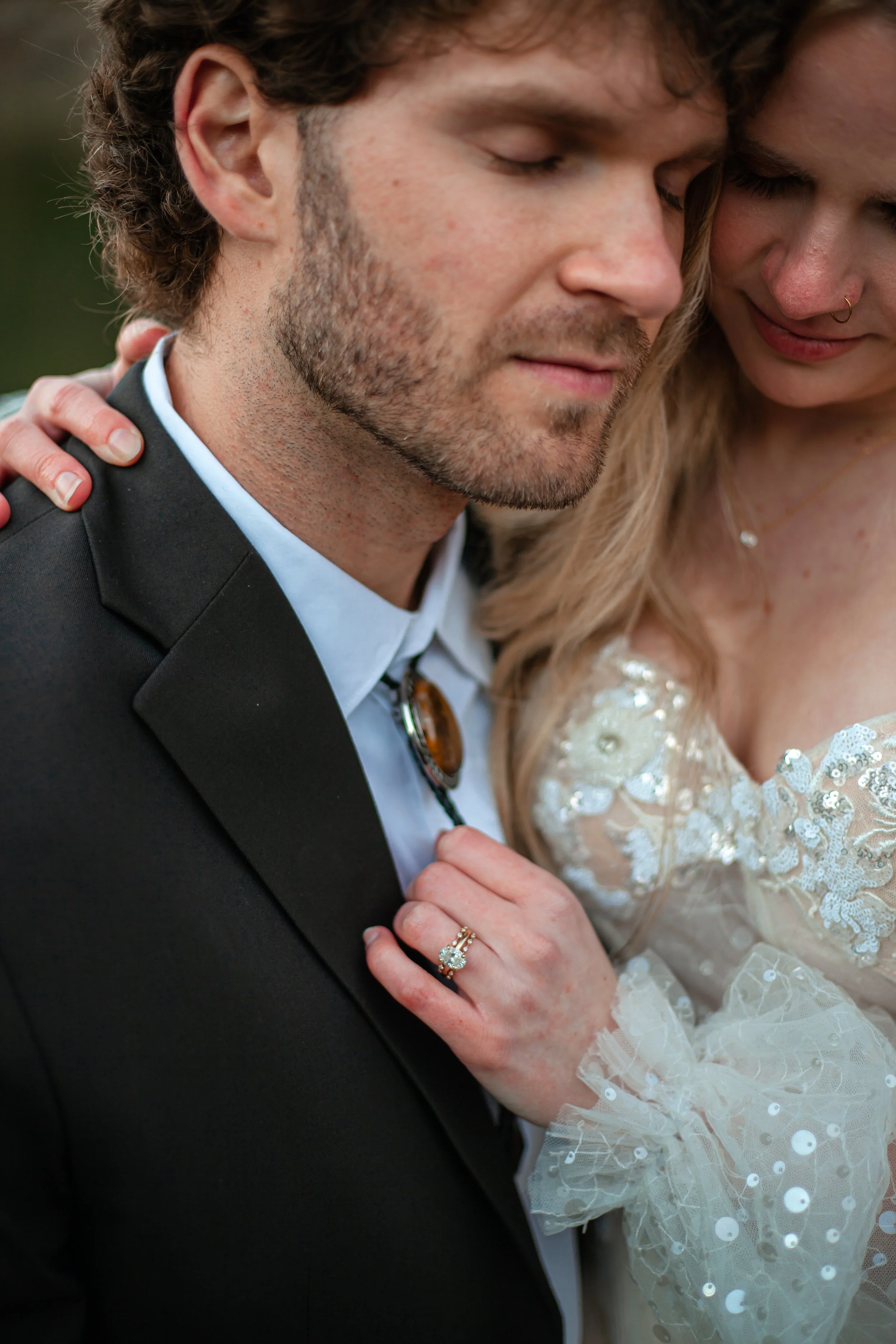 A close-up of a couple, the woman with blonde hair and wearing a lace wedding dress, placing her hand with a wedding ring on the man's chest. The man has curly hair, a beard, and is wearing a suit.