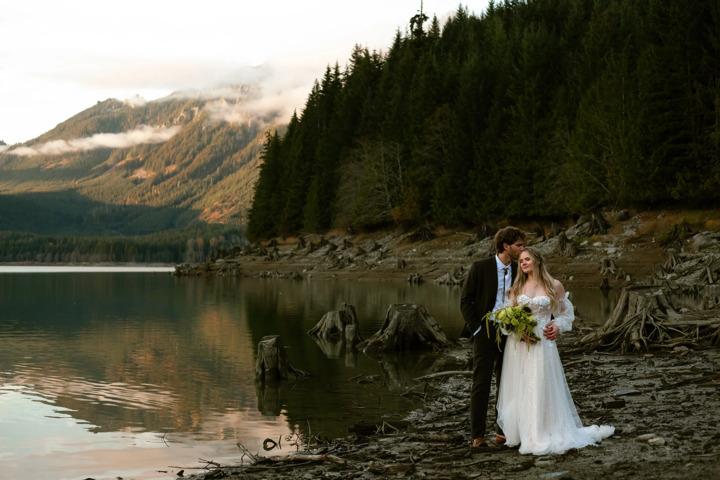 A newlywed couple stands on a rocky lake shoreline surrounded by forested mountains, with clouds and mist in the background. The groom kisses the bride on the forehead as she holds a bouquet of flowers.