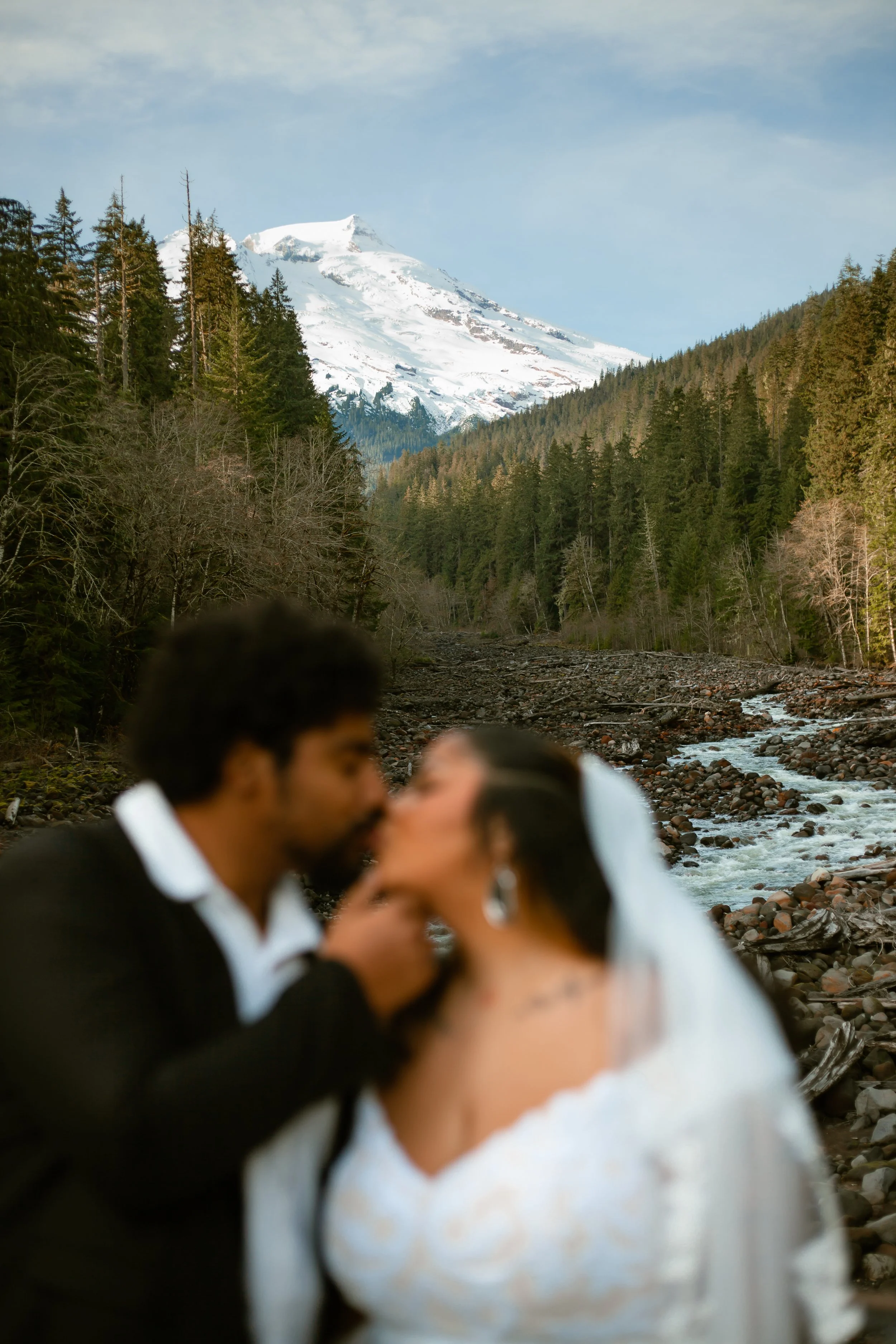 A couple, bride in white wedding dress and groom in black suit, kissing in front of a mountain landscape with snow-capped peaks, evergreen trees, and a flowing river.