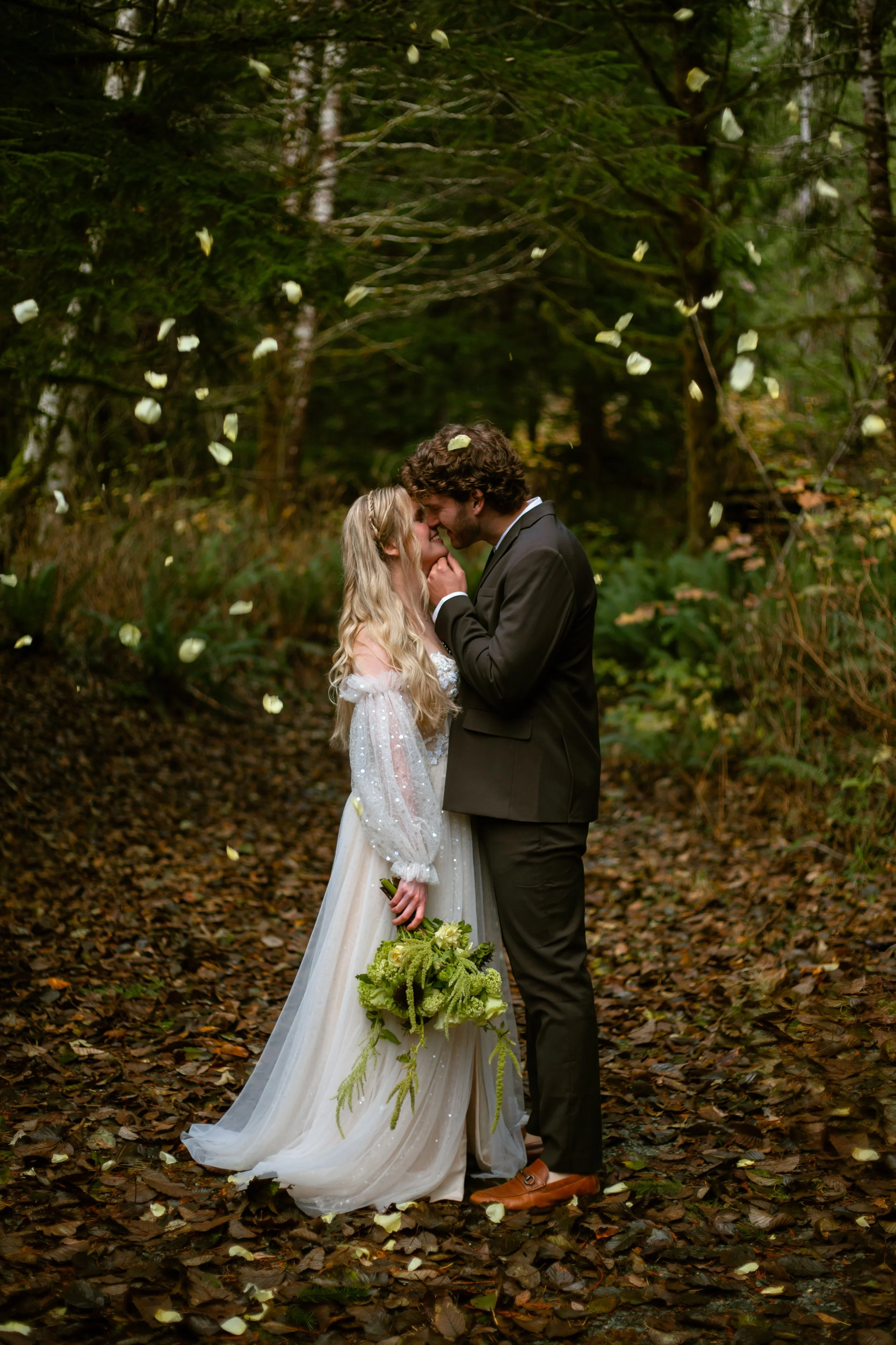 A bride and groom sharing an intimate moment in a forest with falling white petals. The bride is in a white wedding dress holding a bouquet, and the groom is in a black suit. They are touching foreheads, smiling, and standing on a leaf-covered ground