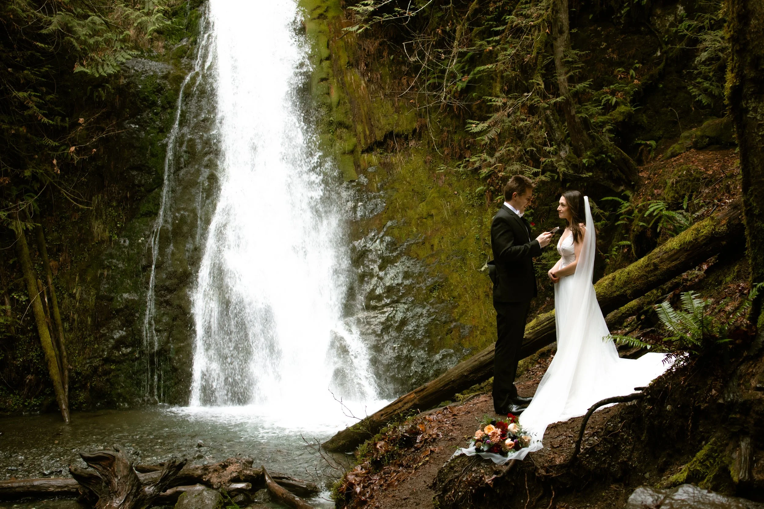 A newlywed couple stands on a rocky lake shoreline surrounded by forested mountains, with clouds and mist in the background. The groom kisses the bride on the forehead as she holds a bouquet of flowers.