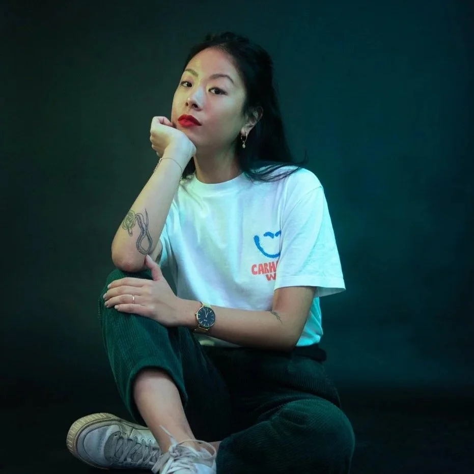 Jeune femme assise, portant un t-shirt blanc avec un dessin de sourire et le texte 'CARH...' en rouge, pantalon noir et baskets blanches, jambes croisées, regardant l'objectif, avec un fond sombre et un éclairage coloré sur le visage