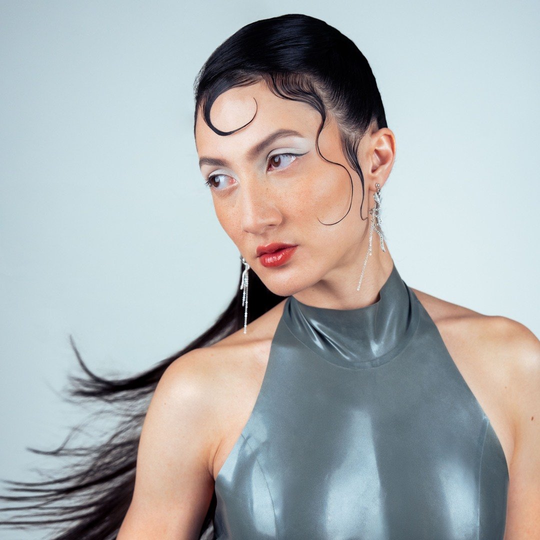 Une femme avec des cheveux noirs lisses et bouclés, portant une robe grise en latex et des boucles d'oreilles longues, pose avec un regard pensif