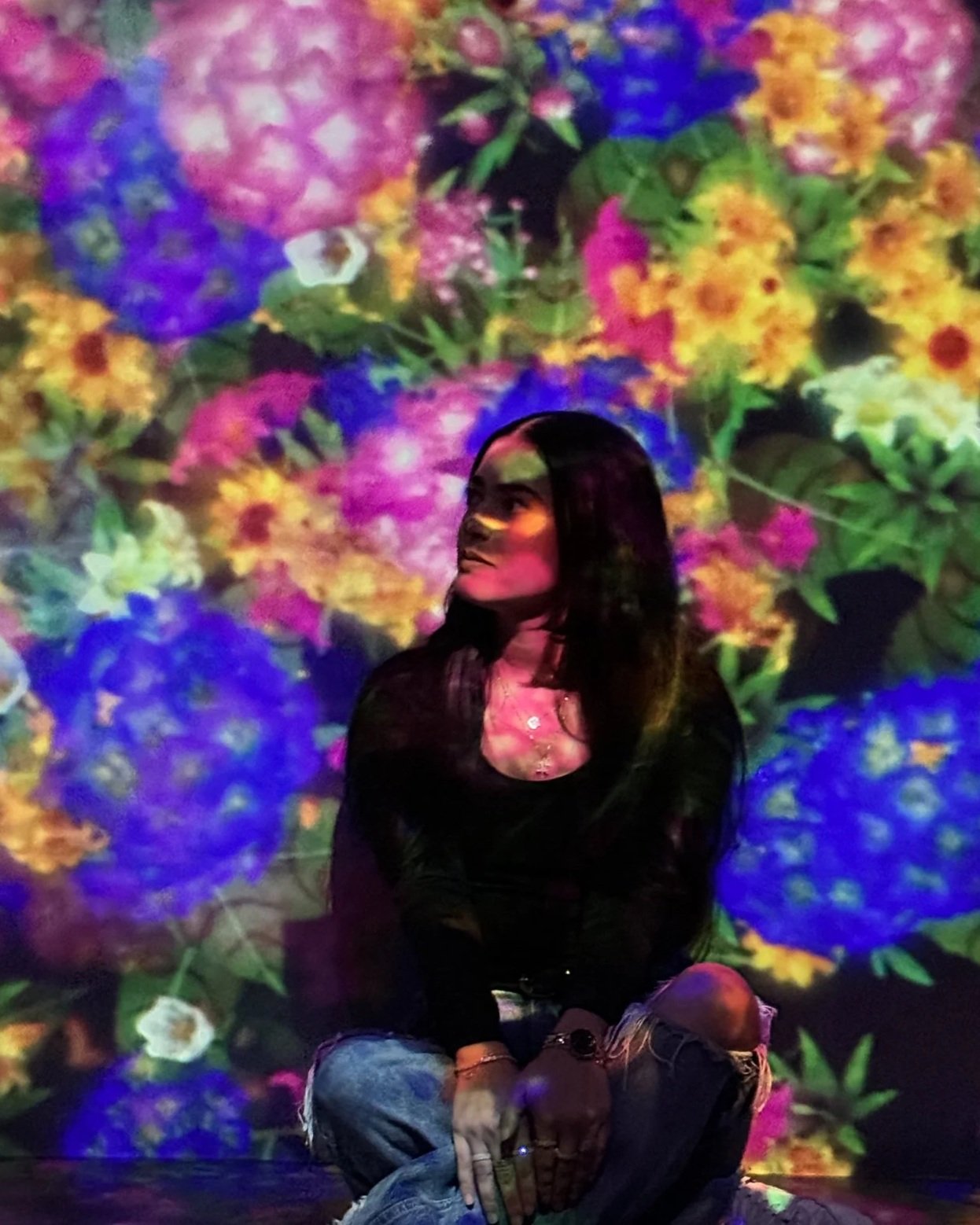 A woman with long dark hair sitting against a vibrant background of colorful flowers projected on a wall or screen.