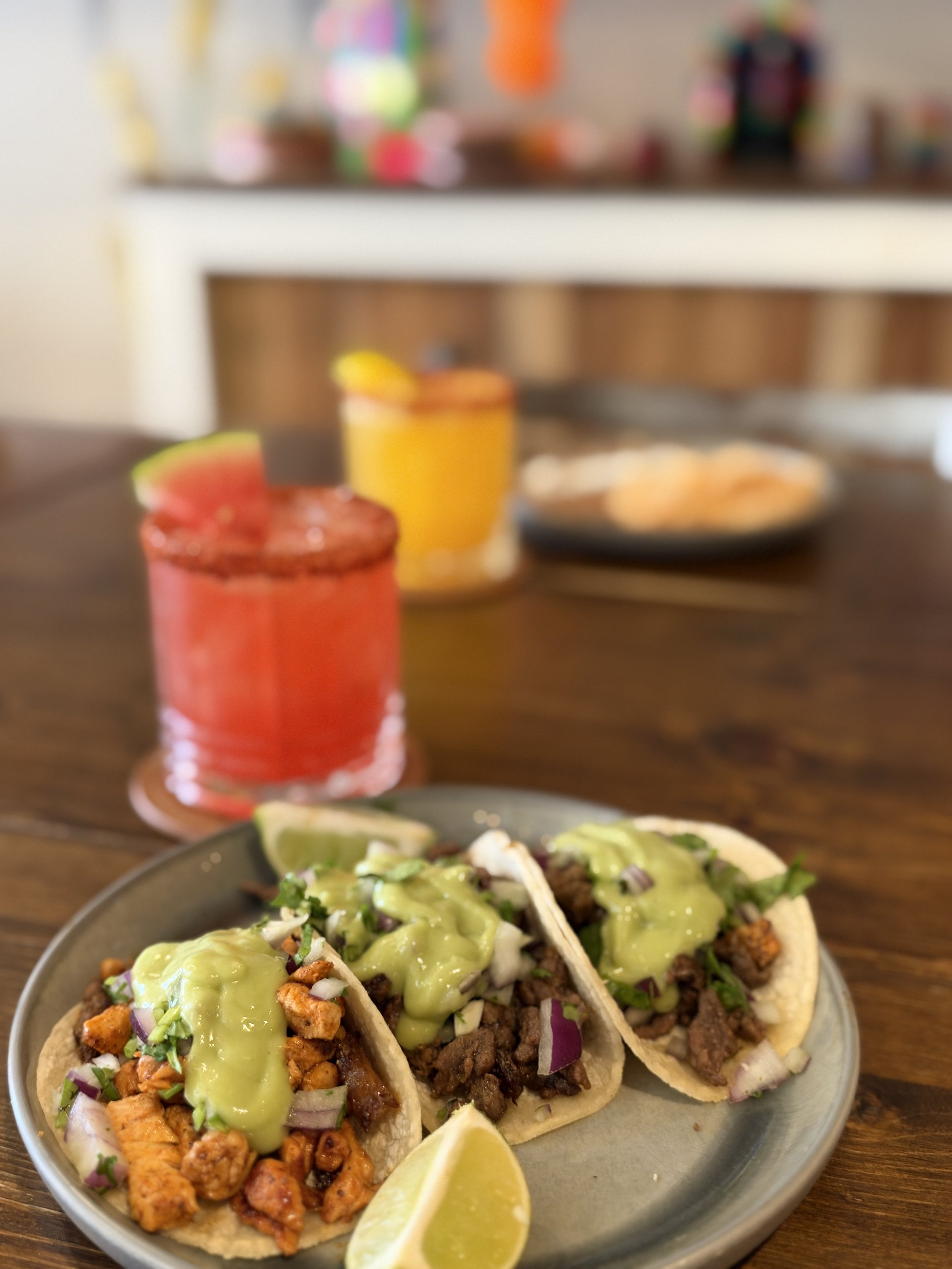 Close-up of three tacos filled with seasoned meat, chopped onions, cilantro, and topped with green sauce, served with lime wedges on a round plate. In the blurred background, there is a table with a drink and a plate of food.