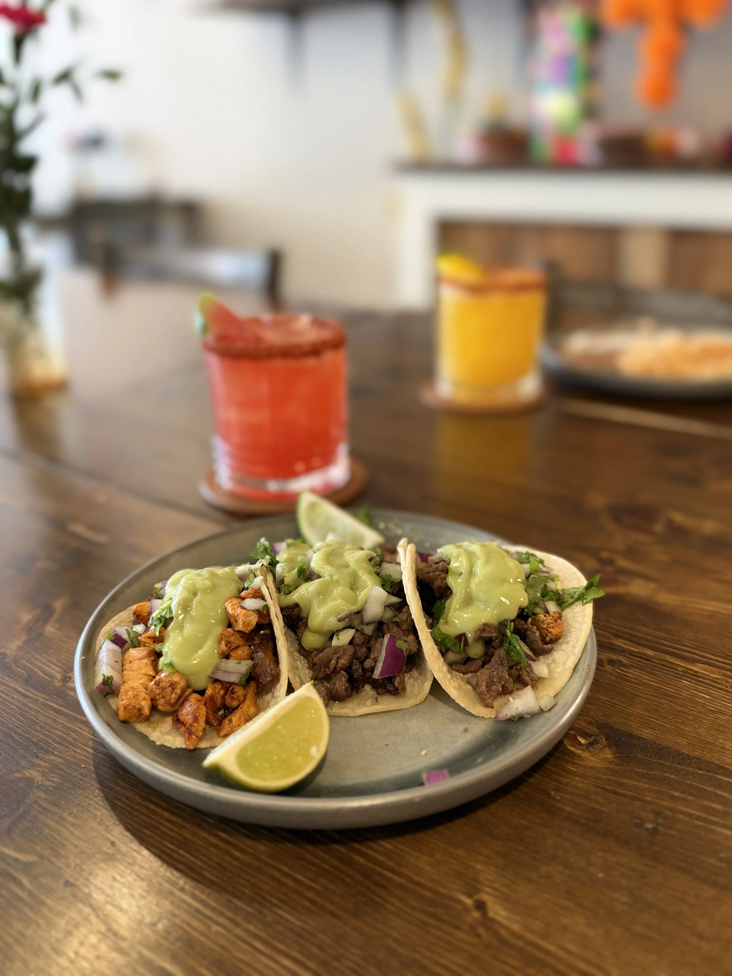 Three street-style tacos with garnishes on a ceramic plate, placed on a wooden table. The tacos are topped with chopped onions, cilantro, and different types of cooked meats, with slices of lime on the side.