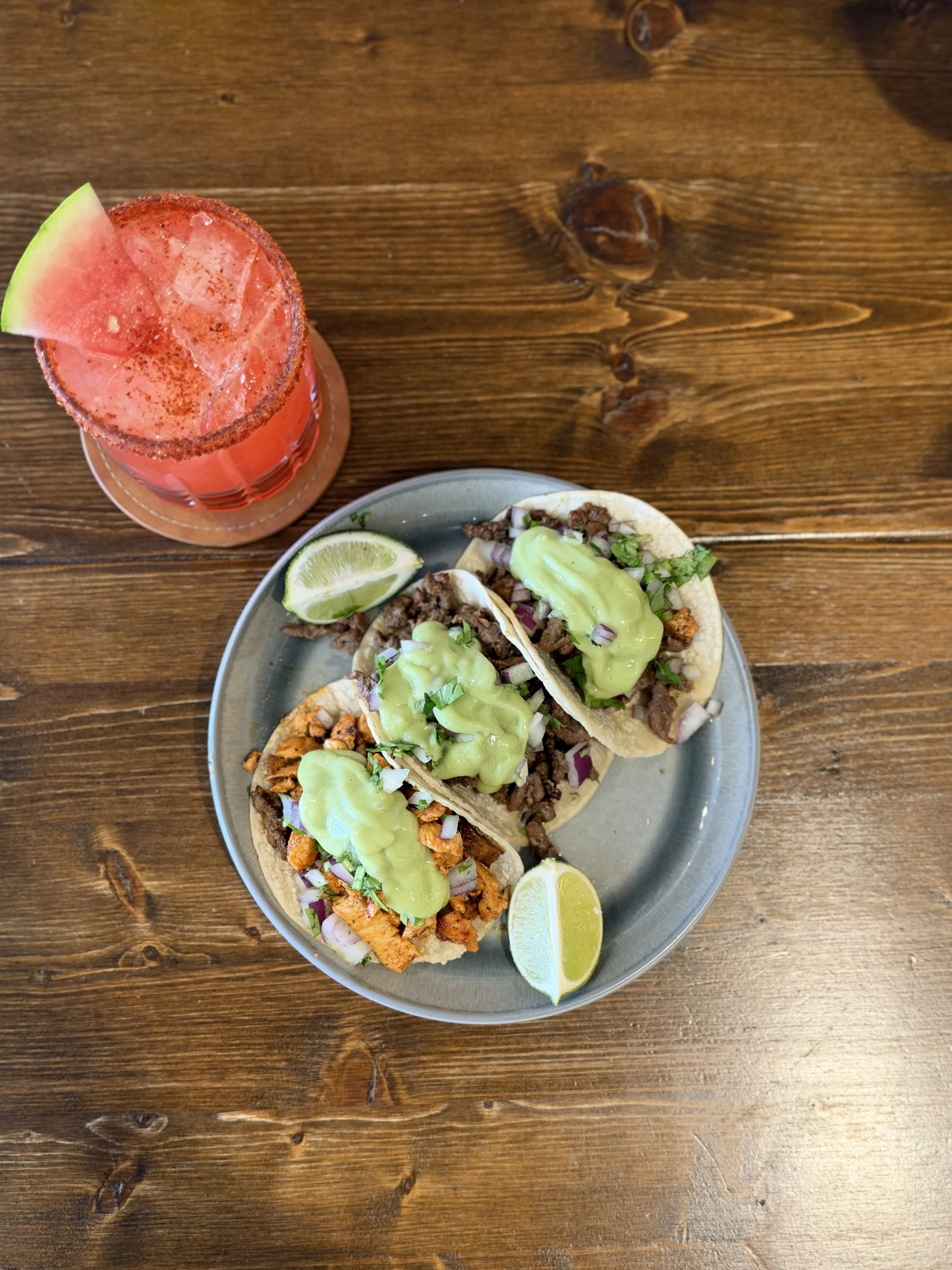 A plate with three tacos filled with meat, vegetables, and sauce, accompanied by lemon wedges, and a red cocktail garnished with a watermelon slice on a wooden table.