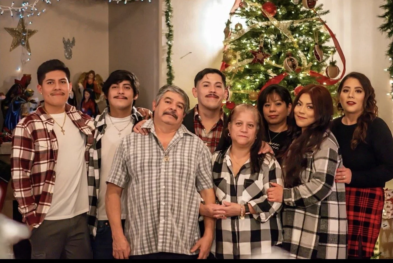 A family gathering in front of a decorated Christmas tree with ornaments, ribbons, and lights. The family members are dressed casually, many in plaid shirts, and are smiling or posing for the camera.