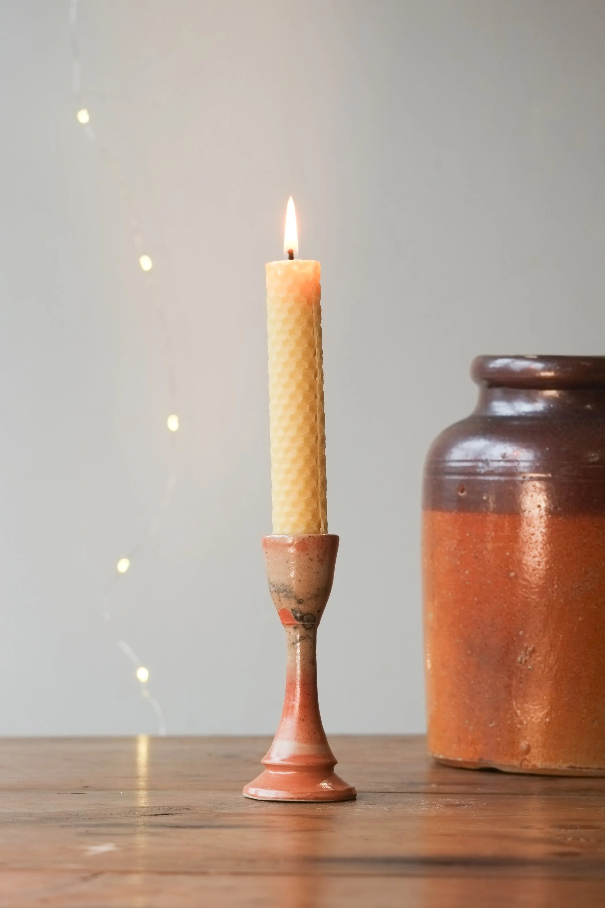A lit beeswax candle in a ceramic holder on a wooden surface with a large ceramic jar to the right and blurred string lights in the background.