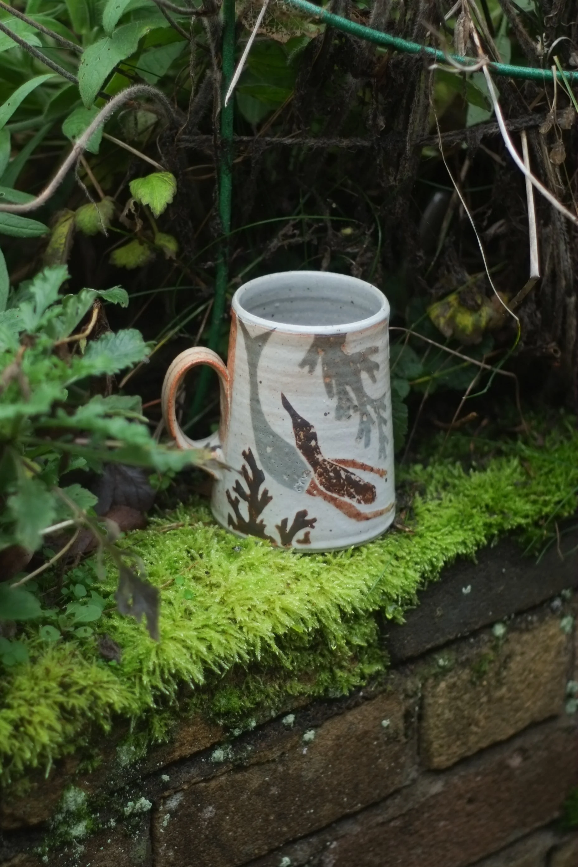 A ceramic mug with leaf and fish designs placed on a mossy brick ledge surrounded by garden plants.