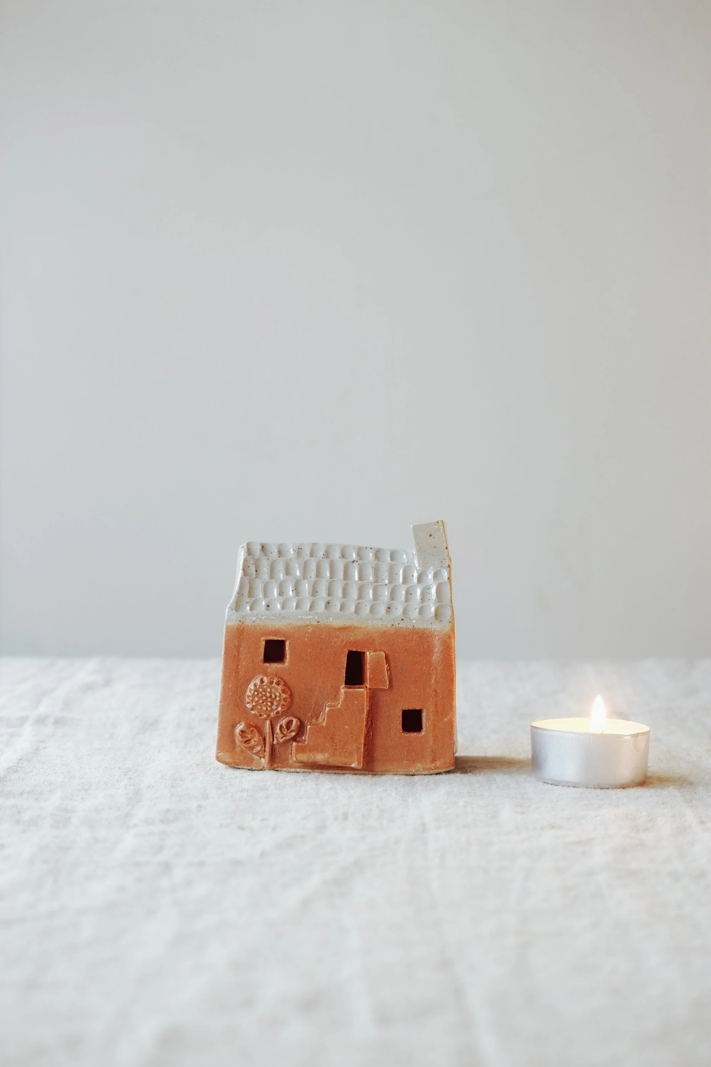 Small ceramic house sculpture with flowers and steps, and a lit tealight candle on a neutral fabric surface against a plain background.