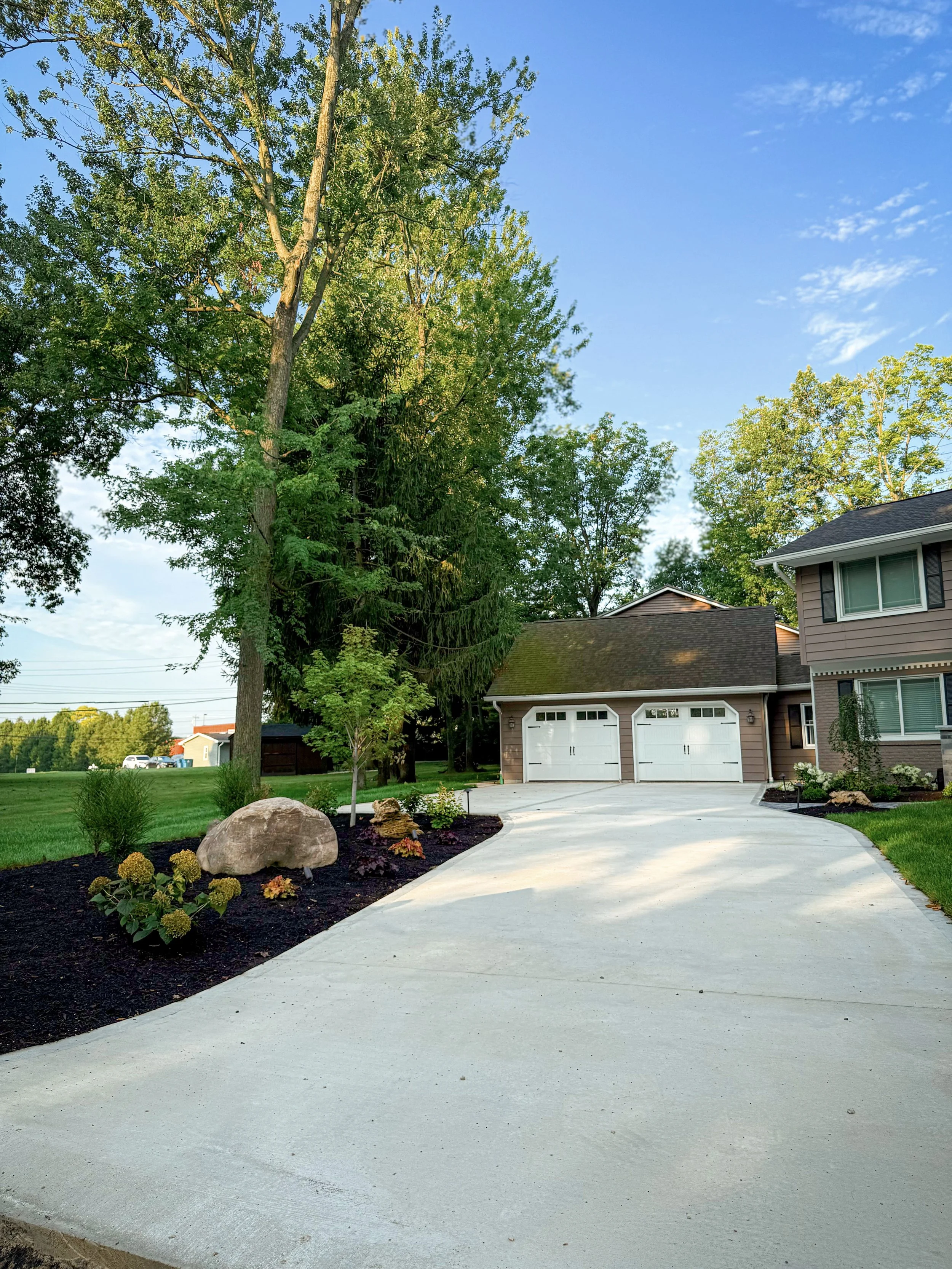 A residential neighborhood with a house, a garage, a driveway, and landscaped yard with grass, trees, rocks, and flowers under a blue sky with some clouds.