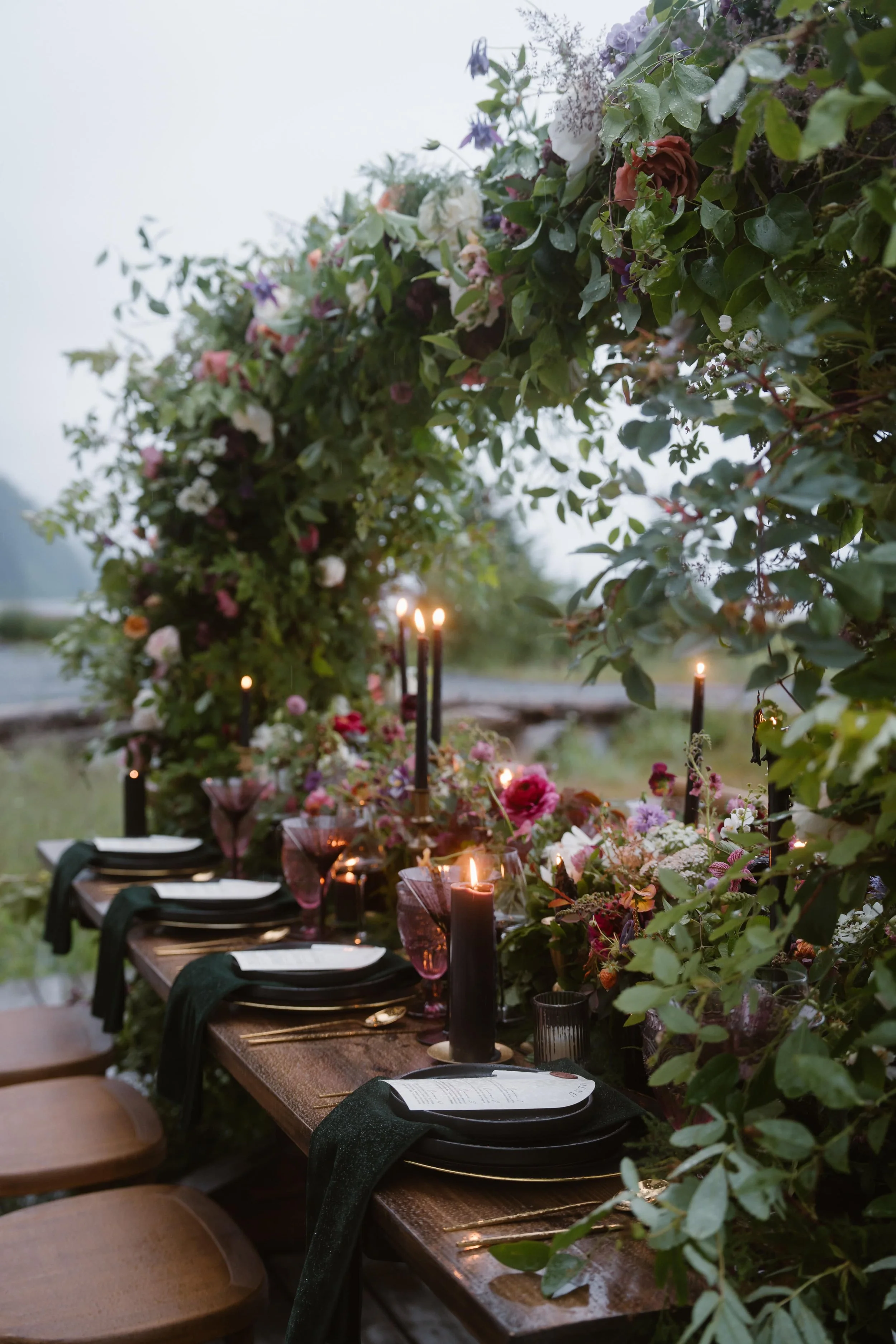 An outdoor dining table decorated with candles, flowers, and place settings under a floral arch.