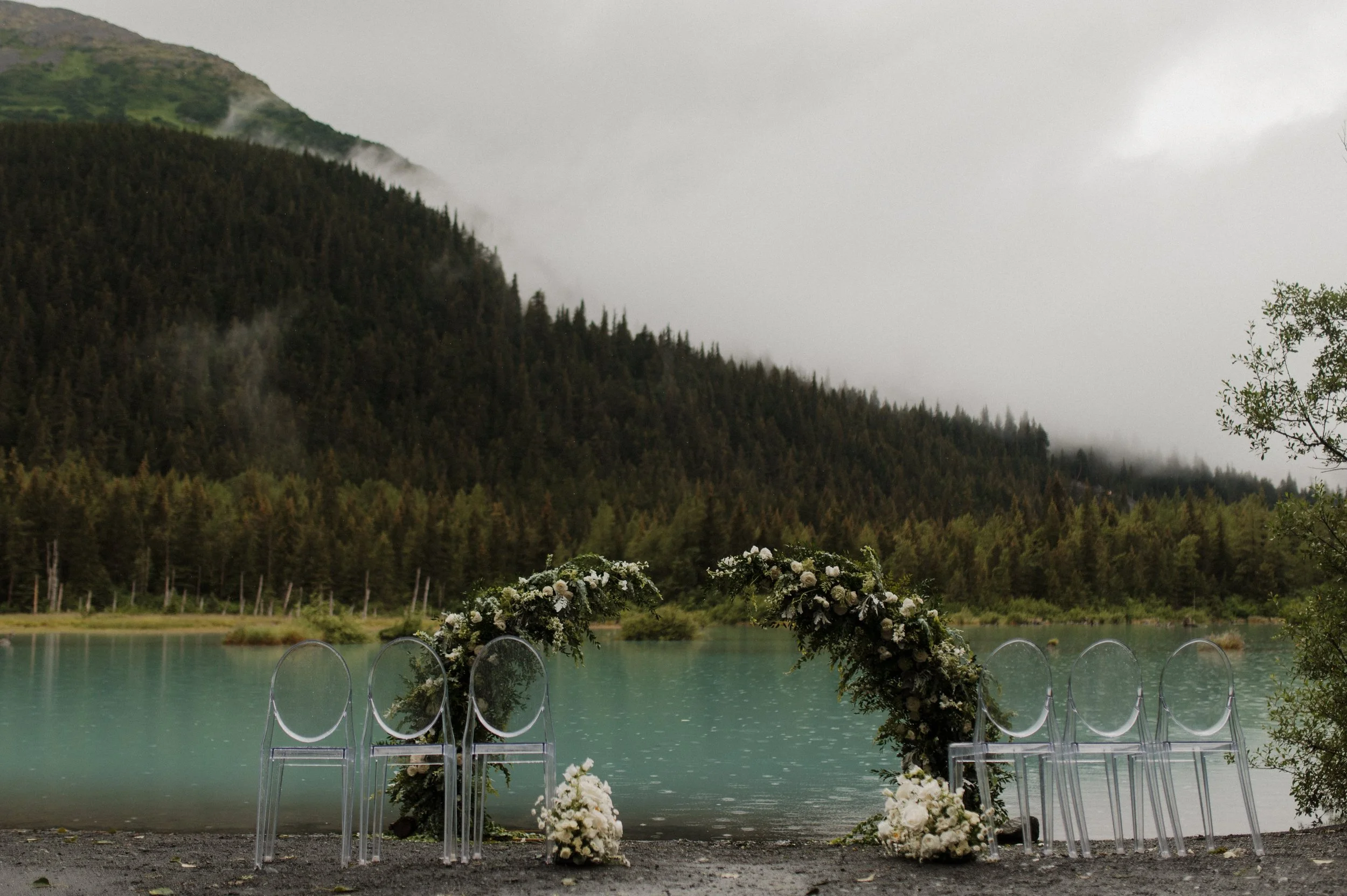 Intimate wedding setup by a lake with a floral arch and white flowers, transparent chairs, on a gray overcast day, with mountains and forest in the background.