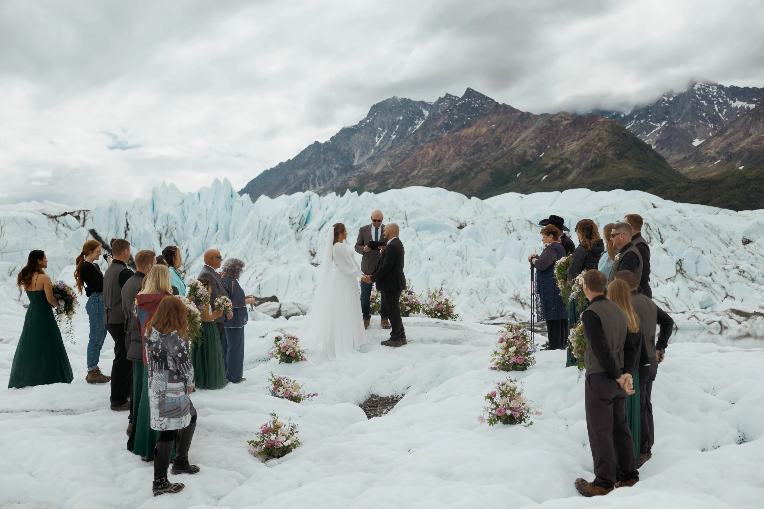 A wedding ceremony taking place on ice in front of a glacier and mountains in a cold, mountainous landscape.