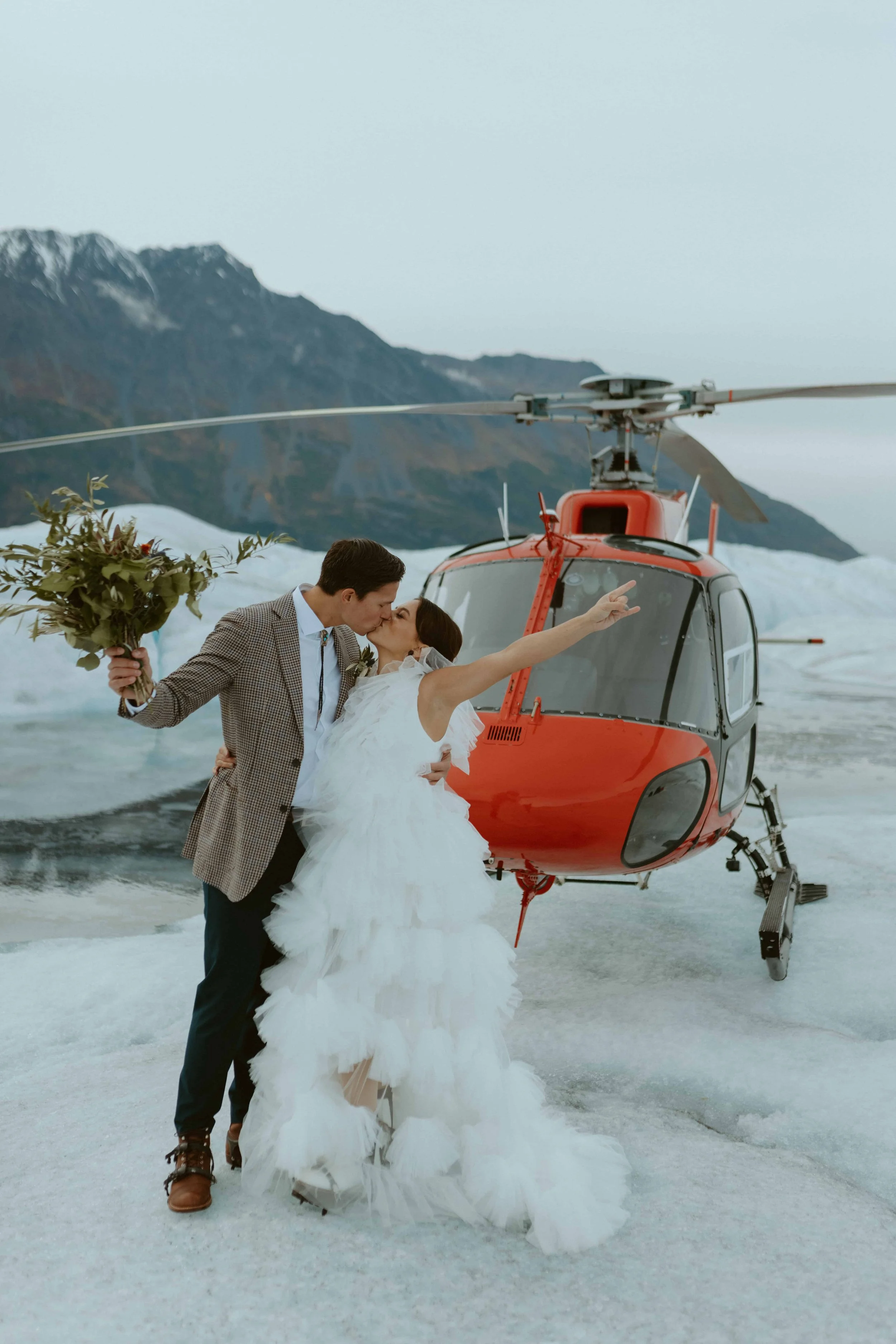 A bride and groom sharing a kiss in front of a red helicopter on an icy landscape, with mountains in the background. The bride is wearing a white wedding gown and holding a bouquet, while the groom is in a checkered jacket and dark trousers.