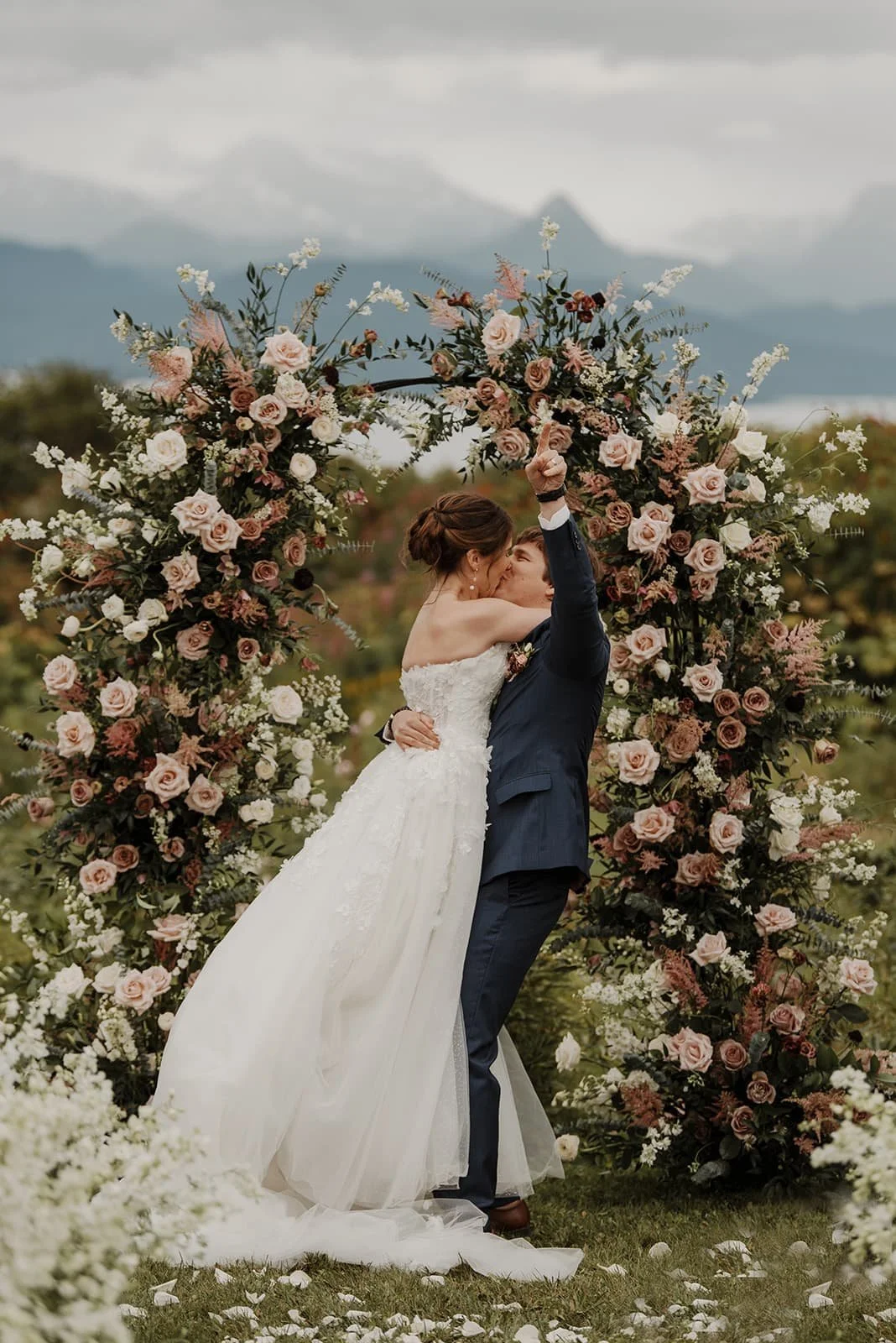 A bride and groom sharing a kiss during their outdoor wedding, surrounded by a large floral arch made of pink and white roses and greenery, with mountains in the background.
