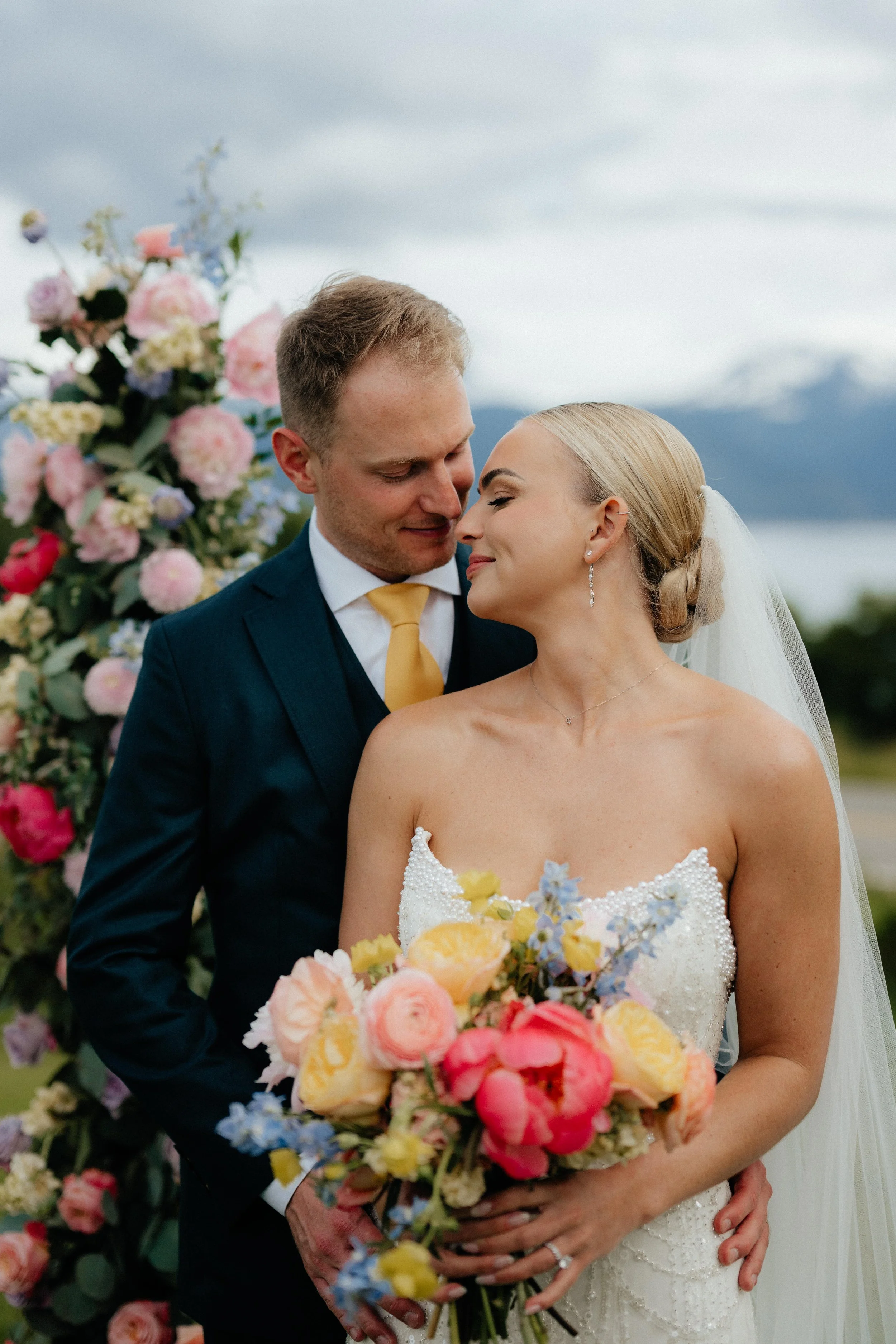 A bride and groom at their wedding, standing close and smiling, with a colorful floral arrangement behind them. The bride holds a large bouquet of pink, yellow, and blue flowers, and wears a white wedding dress with pearl details and a veil. The groo