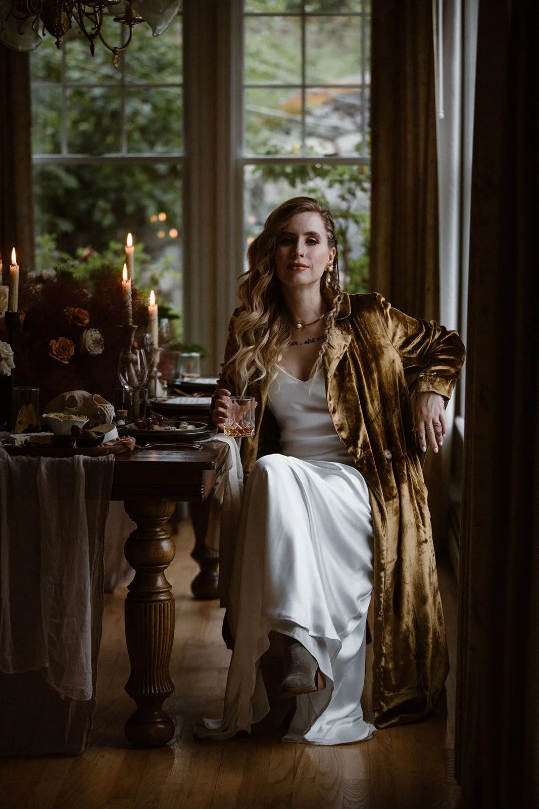 A woman sitting at a dining table holds a glass, wearing a white dress and a long golden brown coat, in a dimly lit room with large windows and decorated with candles and floral arrangements.