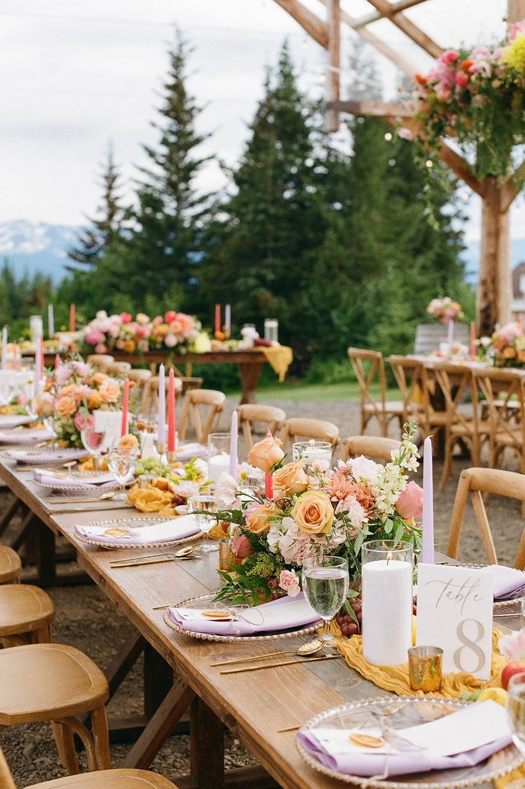 A long outdoor wooden table decorated with floral centerpieces, candles, cutlery, and glasses, set for a wedding reception with a natural mountain and pine forest background.