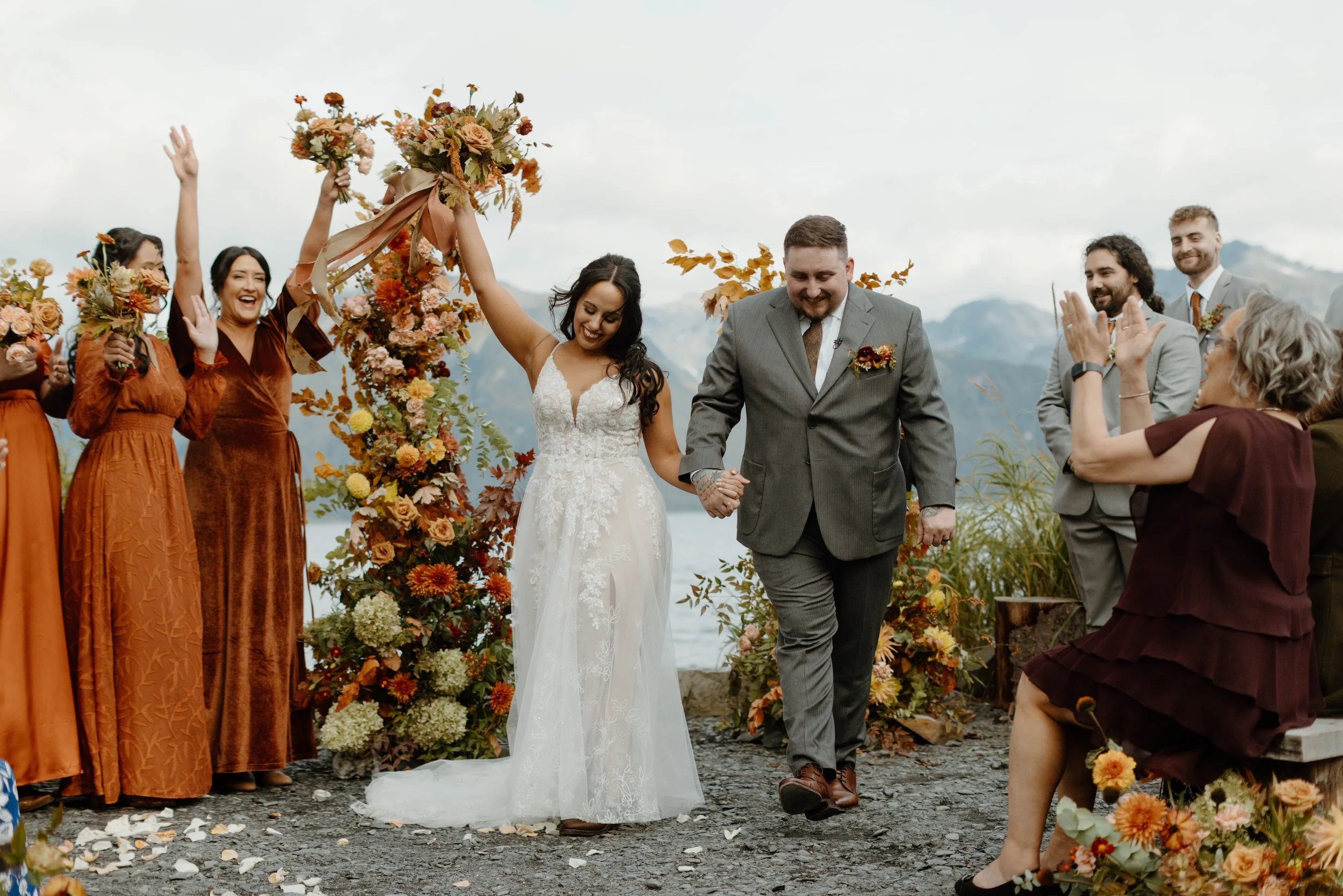 A bride and groom are celebrating their wedding outdoors, holding hands and smiling. The bride is wearing a white lace dress, and the groom is in a gray suit. Bridesmaids and groomsmen, dressed in rust-colored and gray suits, are cheering and clappin