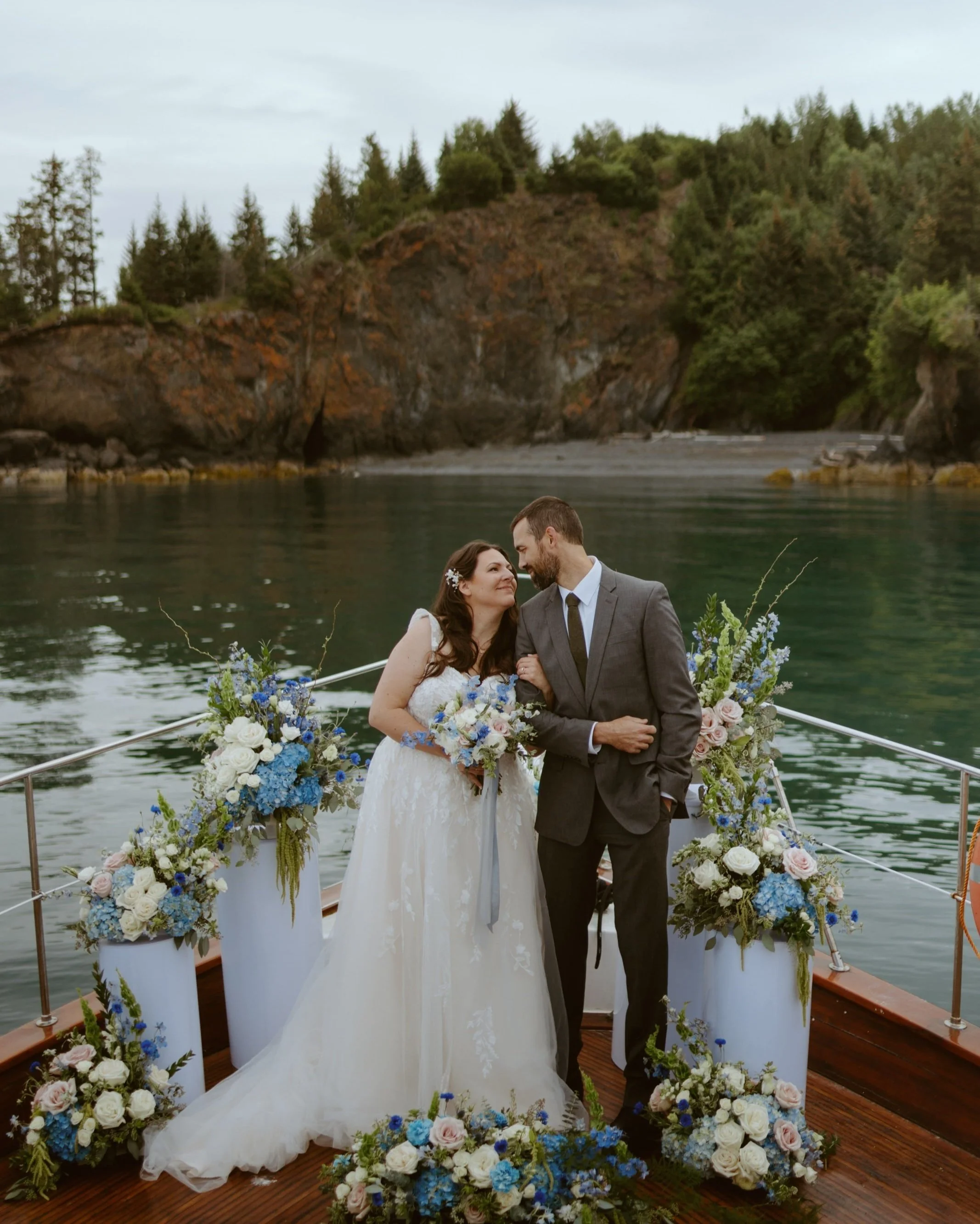 A bride and groom standing on a decorated boat during their wedding ceremony, with ocean and forested cliffs in the background.