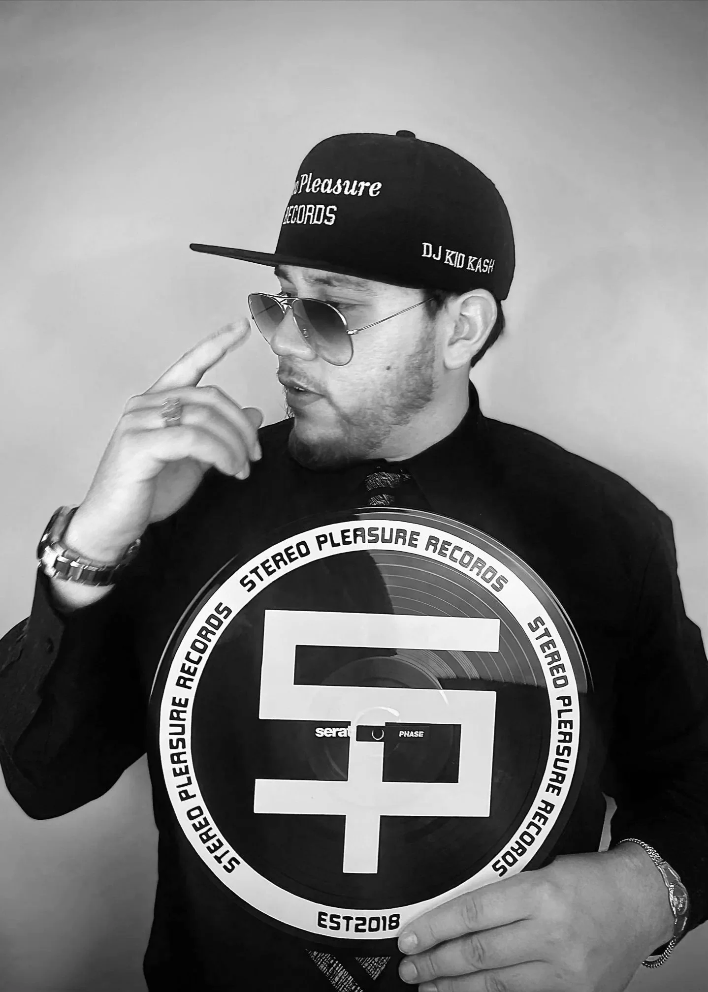 A man wearing sunglasses, a black cap with 'Pleasure Records' and 'DJ Kid Kash' written on it, holding a large record with the Pleasure Records logo, wearing a black shirt and gold watch, standing against a plain background.