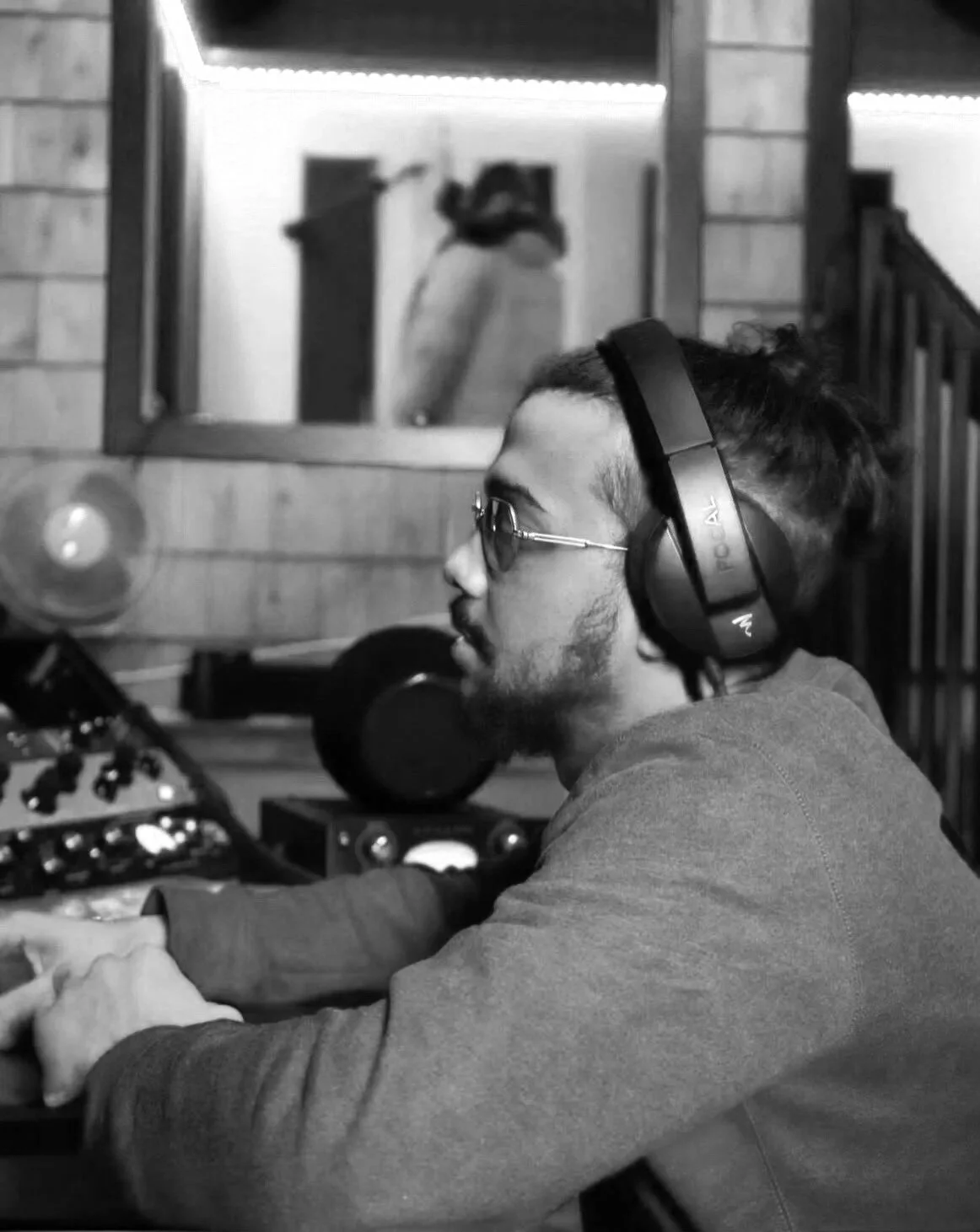 A man with glasses and a beard wearing headphones and a hoodie is operating audio equipment in a recording studio with wooden walls and blue neon lights.