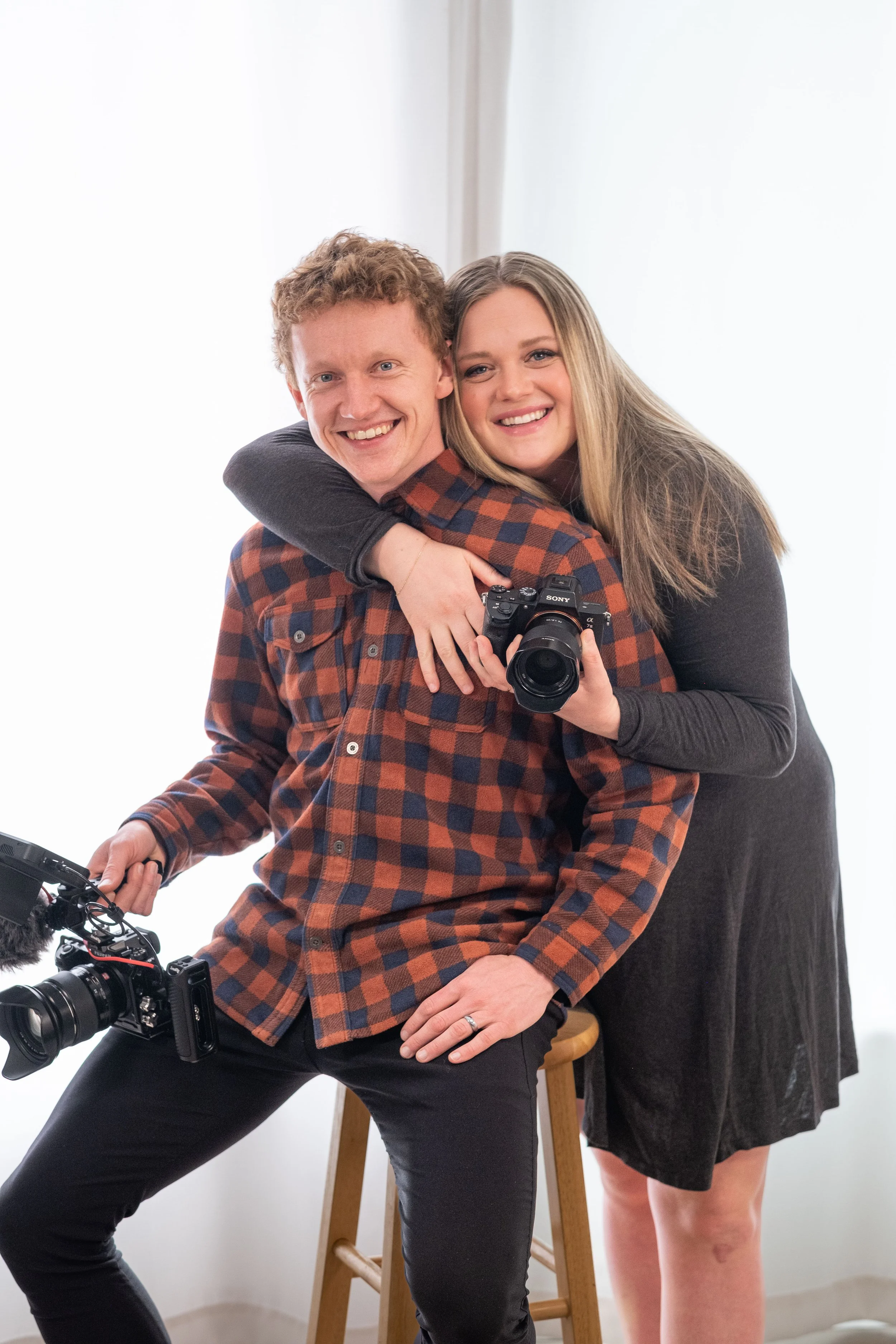 A smiling man and woman posing together, holding cameras, in a bright indoor setting.