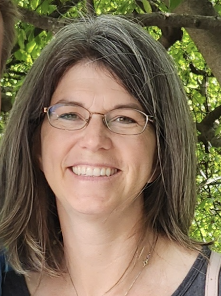 A woman with shoulder-length brown hair, glasses, and a big smile, standing outdoors in front of green foliage and tree branches.