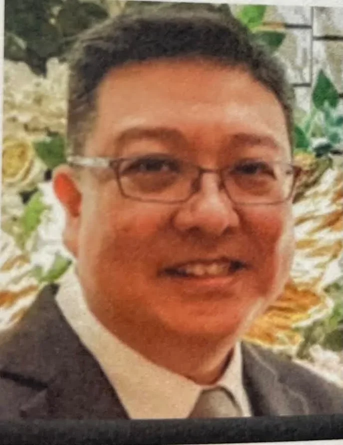 A man with glasses smiling, wearing a suit and tie, standing outdoors with trees and flowers in the background.