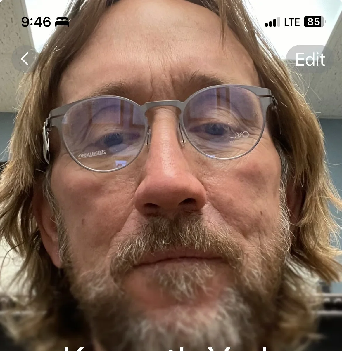 Close-up of a man with glasses and a beard, looking into the camera, with a ceiling and wall in the background.