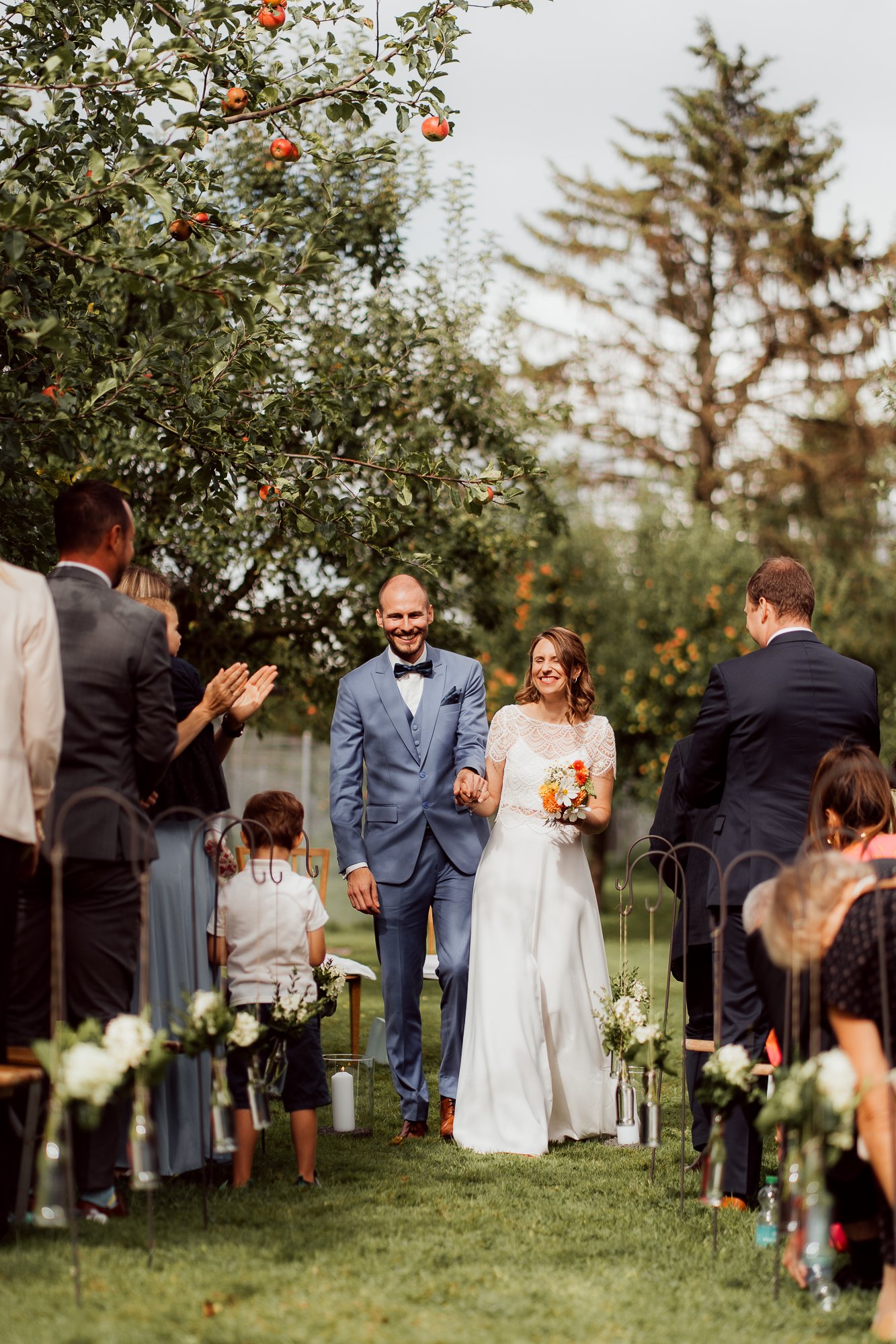 Braut und Bräutigam gehen bei ihrer Hochzeitszeremonie im Freien Hand in Hand, umgeben von Freunden und Familie, unter einem Apfelbaum mit reifen Äpfeln I Hochzeitsfotografie Luzern