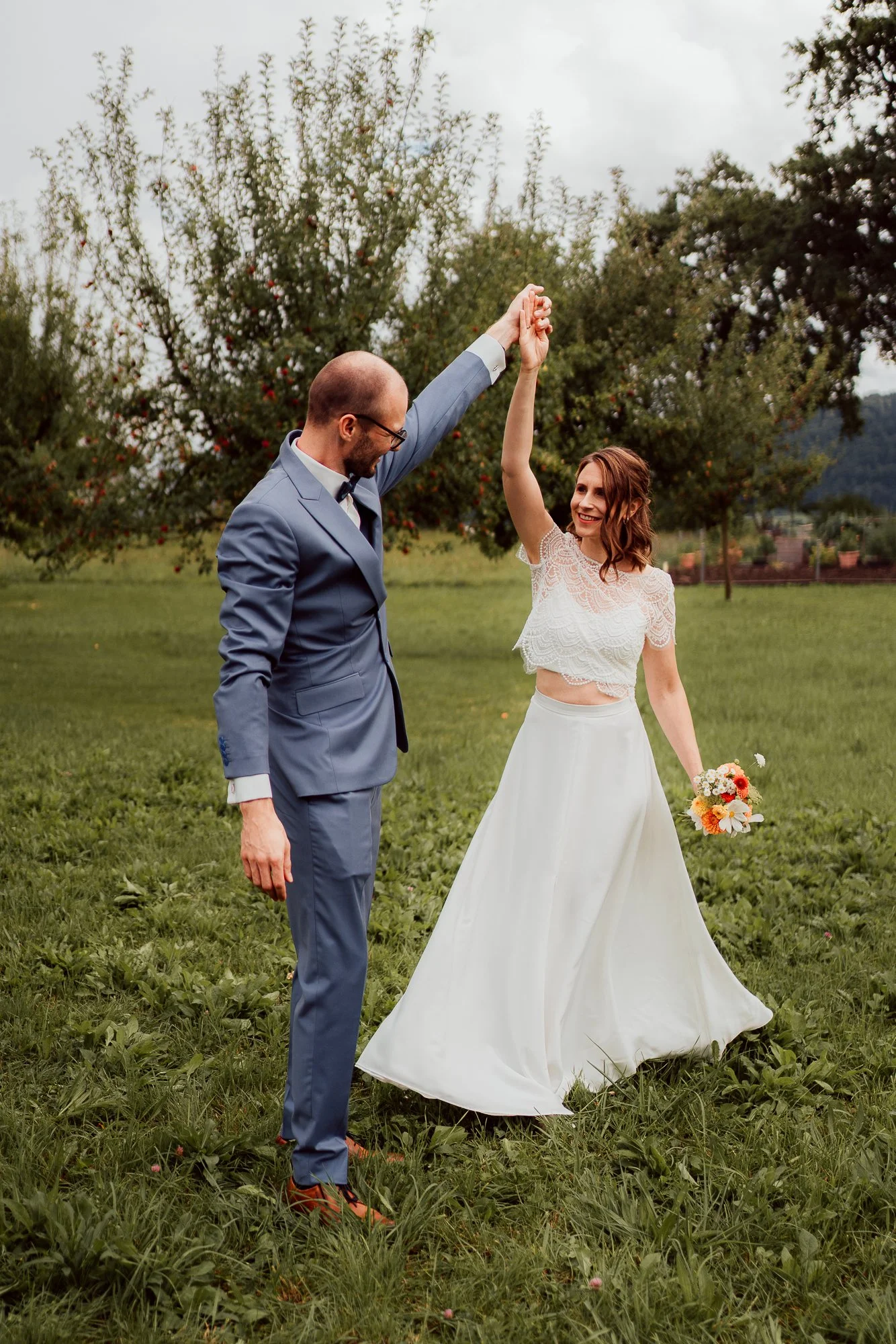 Ein Brautpaar tanzt im Freien, die Frau hält einen Blumenstrauß und lächelt, während der Mann die Hand der Frau hochhält. Im Hintergrund sind Bäume und ein bewölkter Himmel I Hochzeitsfotografie Luzern