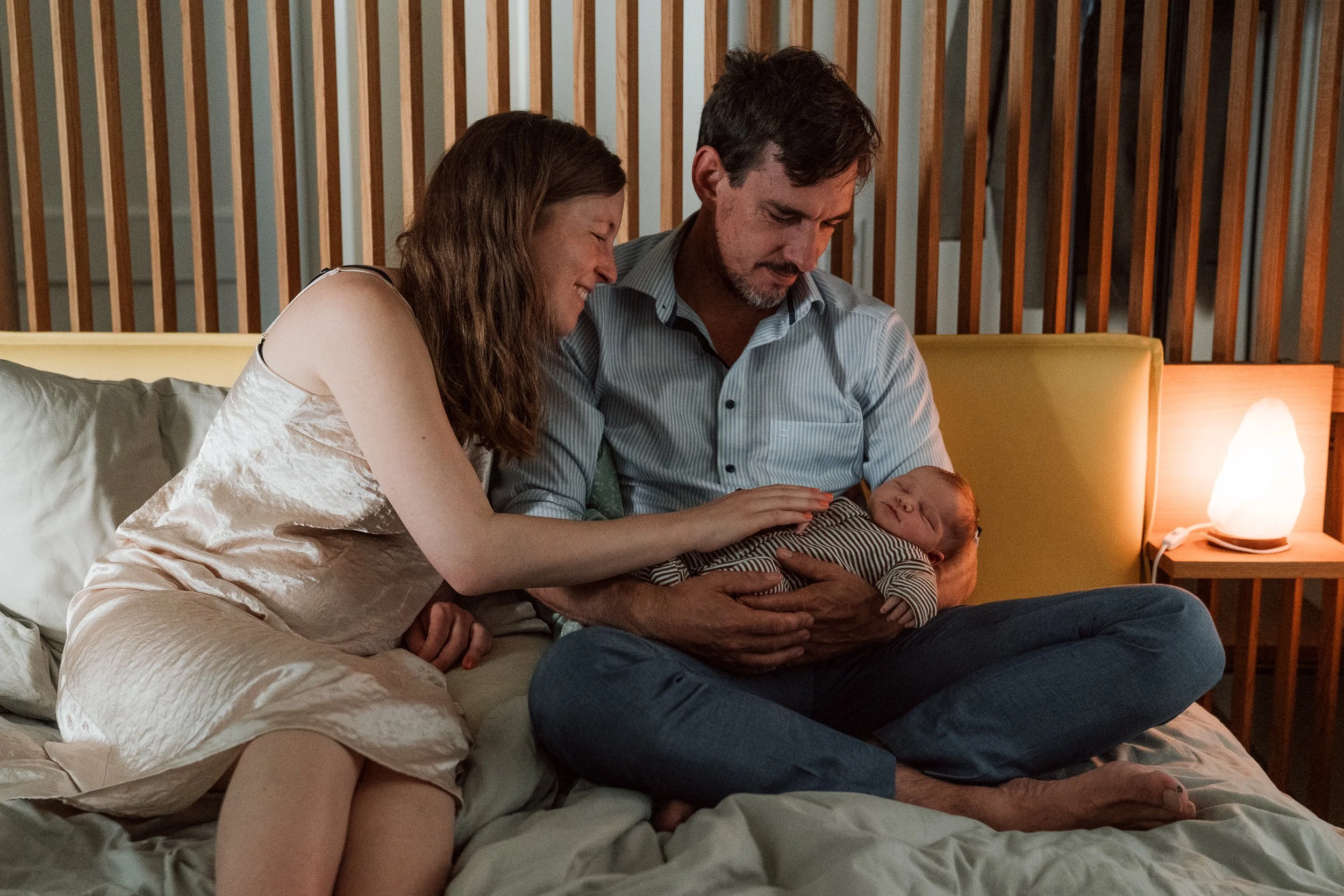 Familie mit Neugeborenem im Bett im Wochenbetthaus Terra Alta, Erwachsene sitzen auf dem Bett, Baby wird gehalten, Lichter im Hintergrund I Wochenbettreportage, Neugeborenenfotografie Luzern