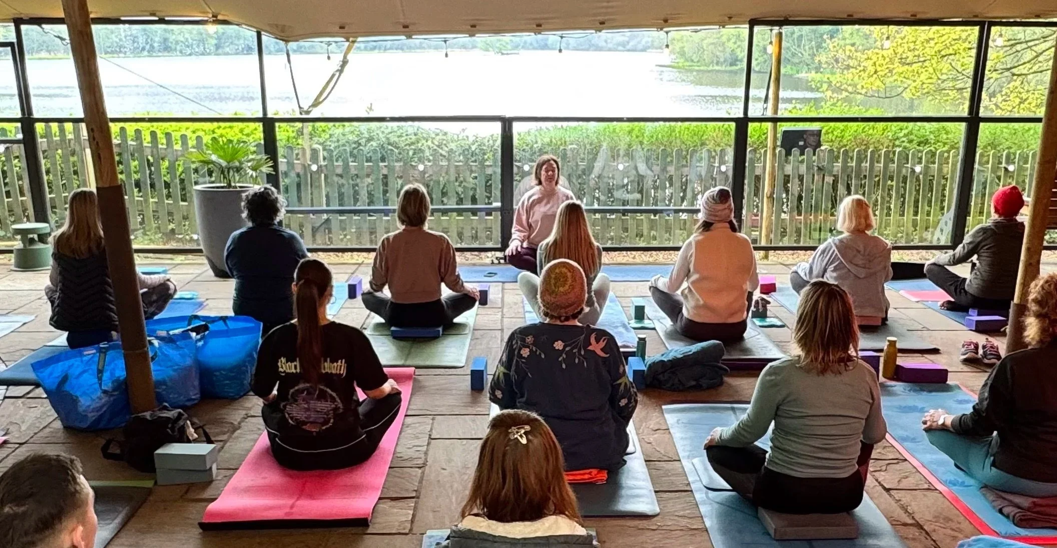 A group of people participating in a yoga class on mats, seated on their knees or crossed-legged, facing an instructor who is demonstrating meditation or breathing exercises. They are inside a wooden and glass structure overlooking a scenic outdoor area with a lake or river and trees.