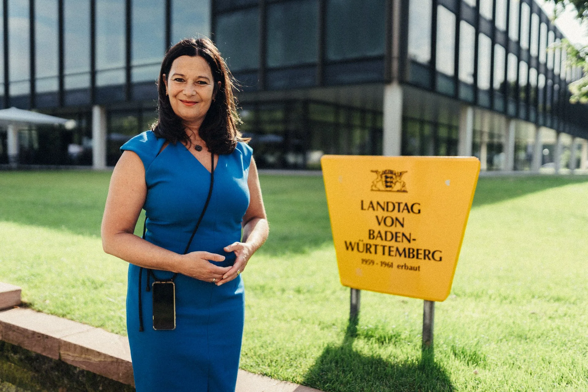 Katrin Steinhülb-Joos steht in einem blauen Sommerkleid vor dem Landtagsgebäude. Neben ihr steht ein Hinweisschild mit der Aufschrift "Landtag von Baden-Württemberg, 1959 bis 1961 erbaut"