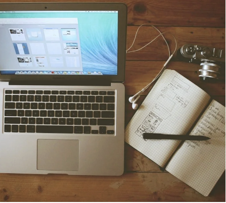 Open laptop on a wooden table with a photograph editing application on the screen, next to a notebook with sketches and notes, a black pen, a camera, and a white charging cable.