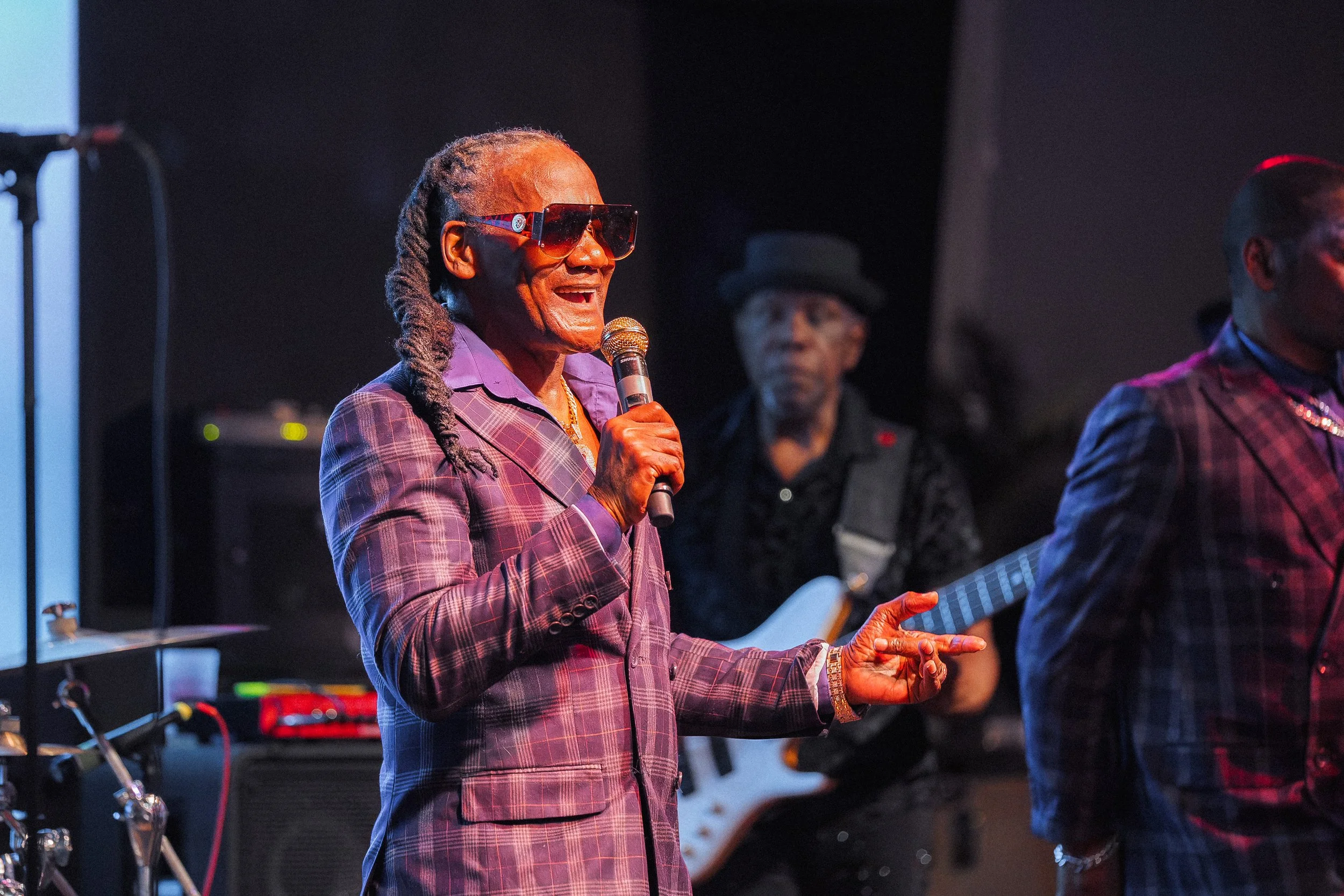 An elderly man with long gray dreadlocks, wearing large sunglasses and a purple plaid suit, singing into a microphone on stage with a band playing behind him.