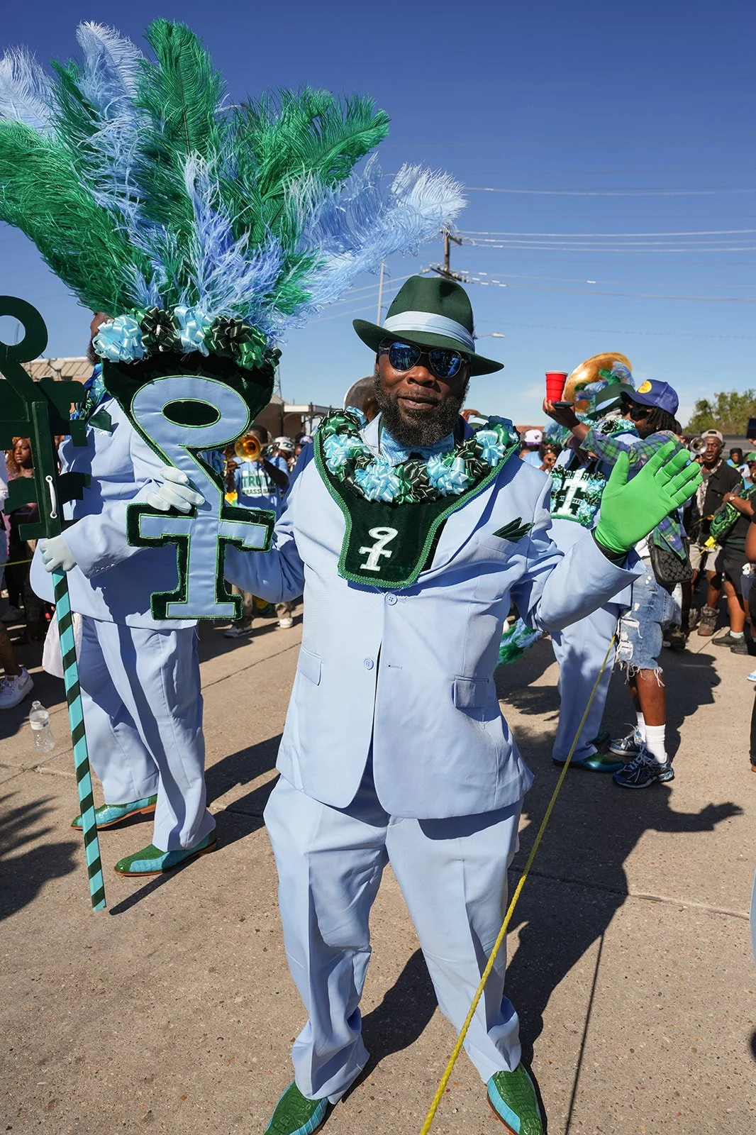 A man dressed in a white suit and green accessories at a parade, holding a large symbol and wearing a feathered headpiece, surrounded by other parade participants.