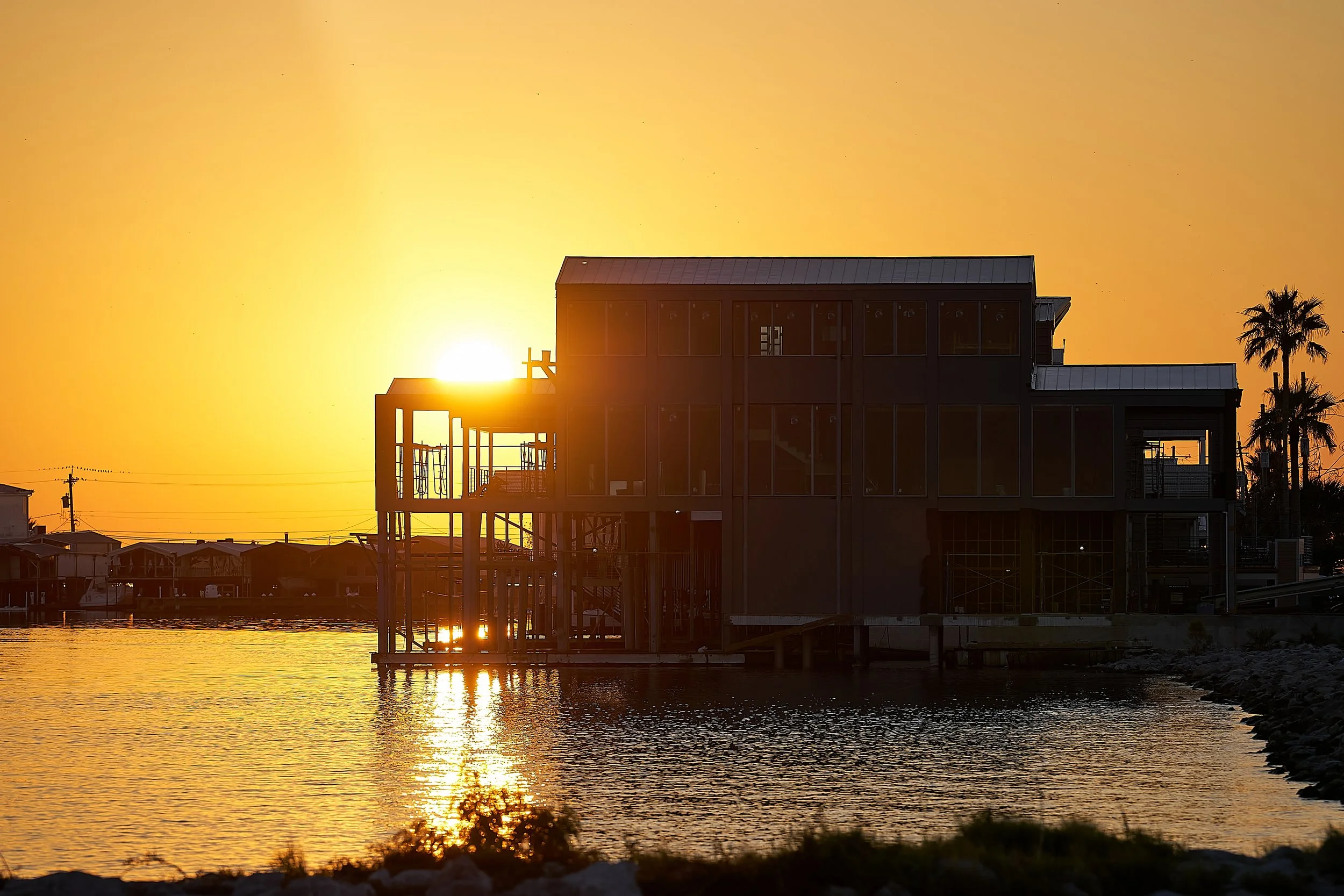 Sunset over a modern house built on stilts by the water, with the sun partially visible behind the structure, orange sky, and palm trees in the background.