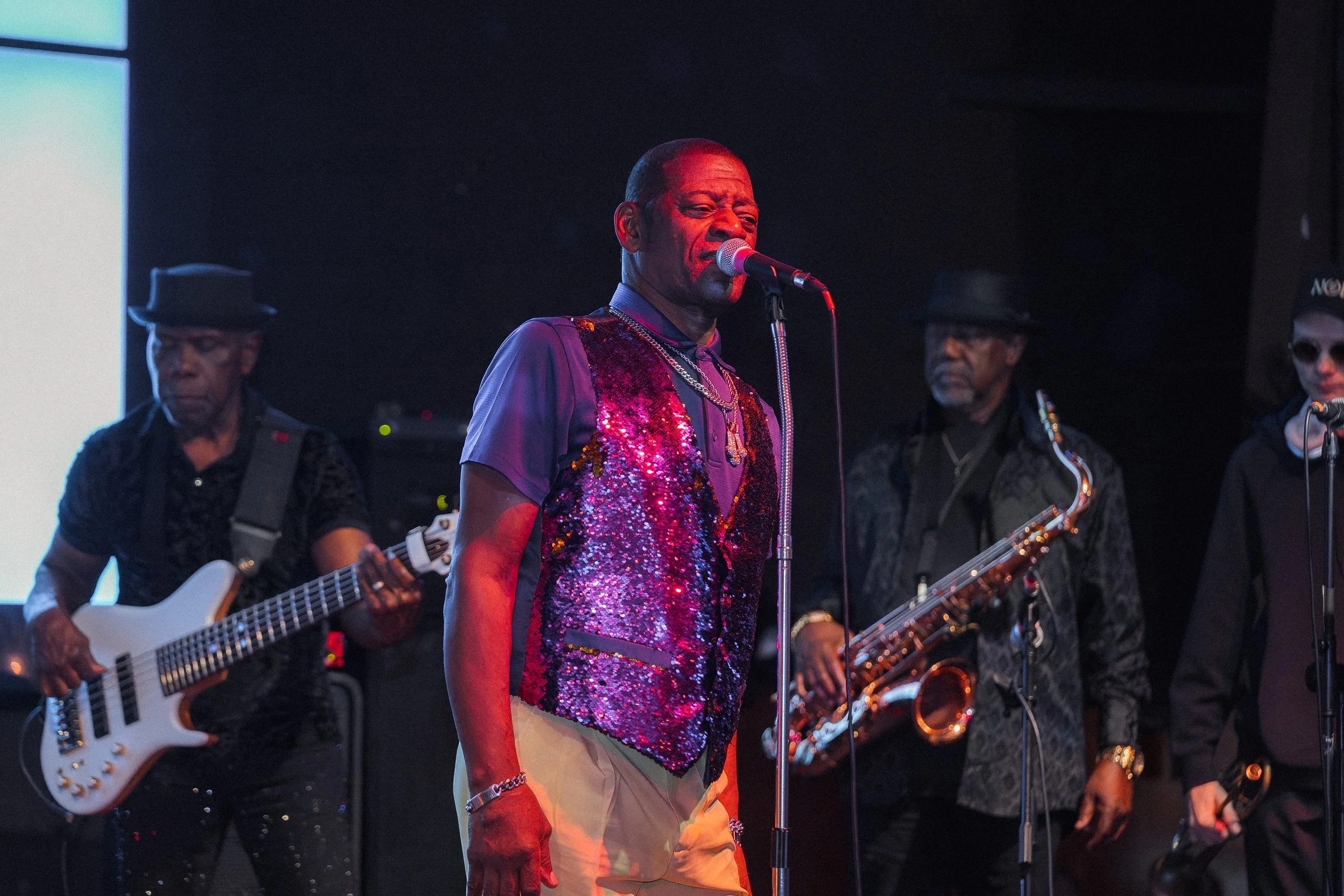 Male singer in a colorful sequin vest performing on stage with musicians playing guitar and saxophone in the background.
