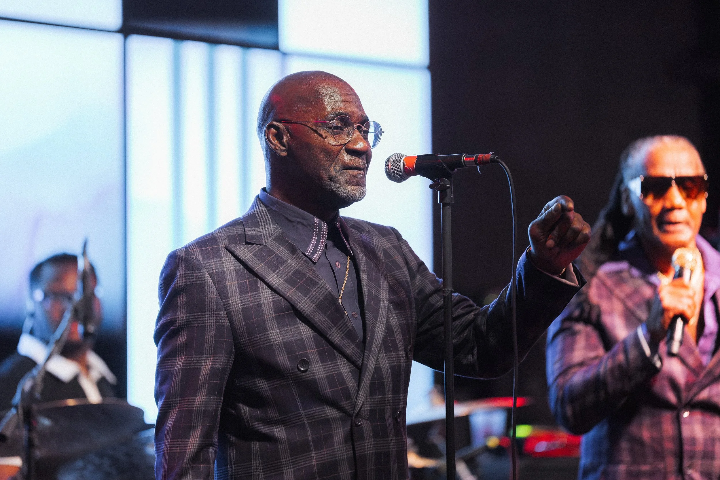 An older man wearing glasses and a plaid suit standing in front of a microphone during a performance or speech, with a woman and a man in the background also holding microphones, on a stage with large windows or light panels behind them.