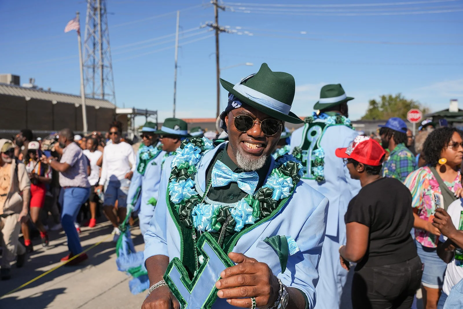 A man smiling and wearing sunglasses, a green hat, a light blue suit, a blue bow tie, and decorated with leis, in a crowd during a lively outdoor celebration or parade.
