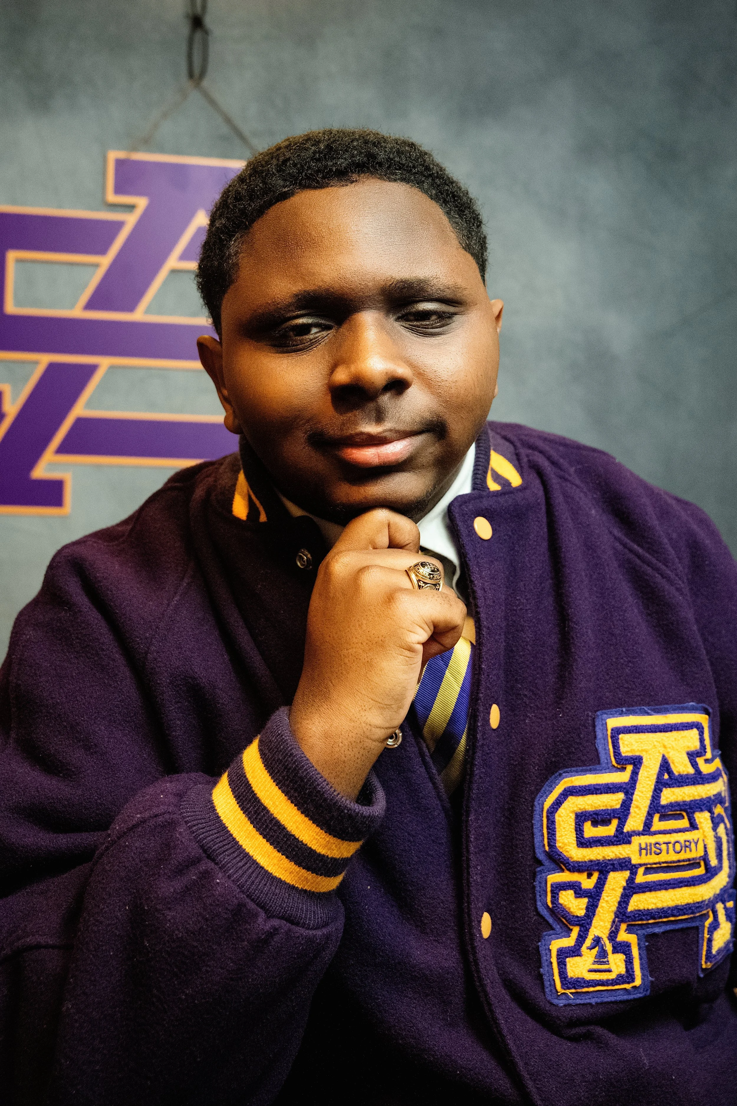 Young man with dark skin wearing a purple jacket with a yellow and purple emblem that says 'HISTORY.' He is holding his chin with his right hand and looking at the camera. The background features a purple and yellow logo on a gray wall.