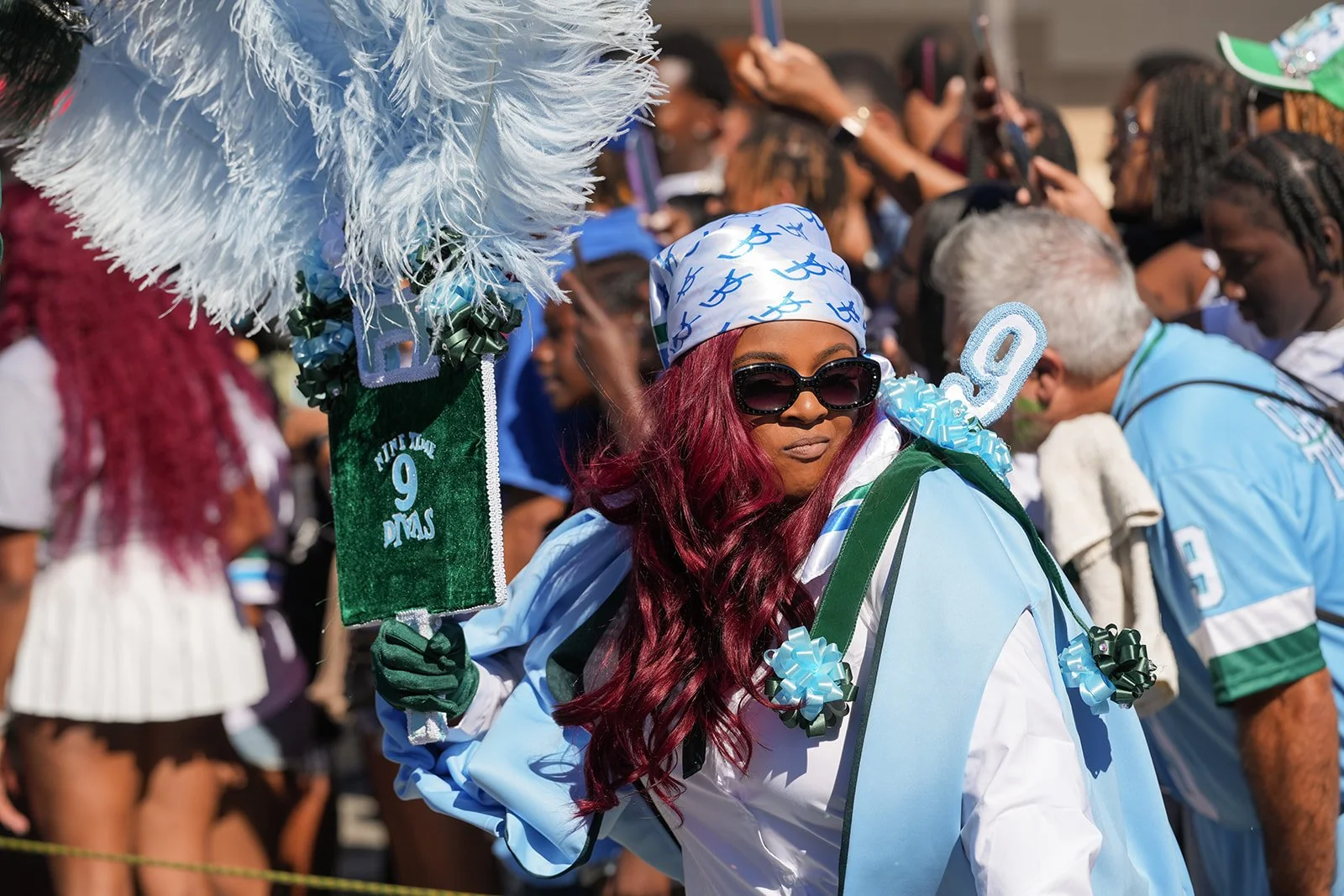 Woman with long burgundy hair wearing oversized sunglasses, a blue and white bandana, and a light blue jacket, holding a green and white pom-pom fan, at a crowded outdoor event or parade.
