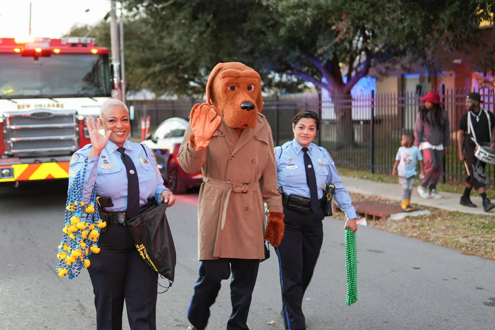 Police officers and a person in a dog costume walking on a street during a parade or celebration, with a fire truck and onlookers in the background.