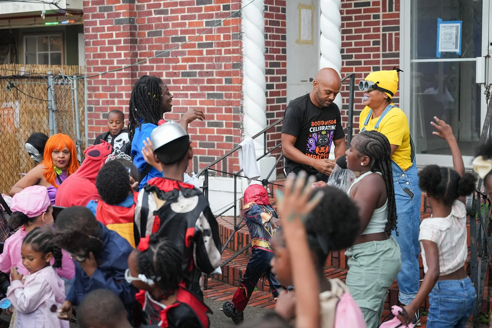 A diverse group of children and adults celebrating outdoors, many dressed in costumes, with a brick house in the background. The scene appears festive with people talking, laughing, and engaging with each other.